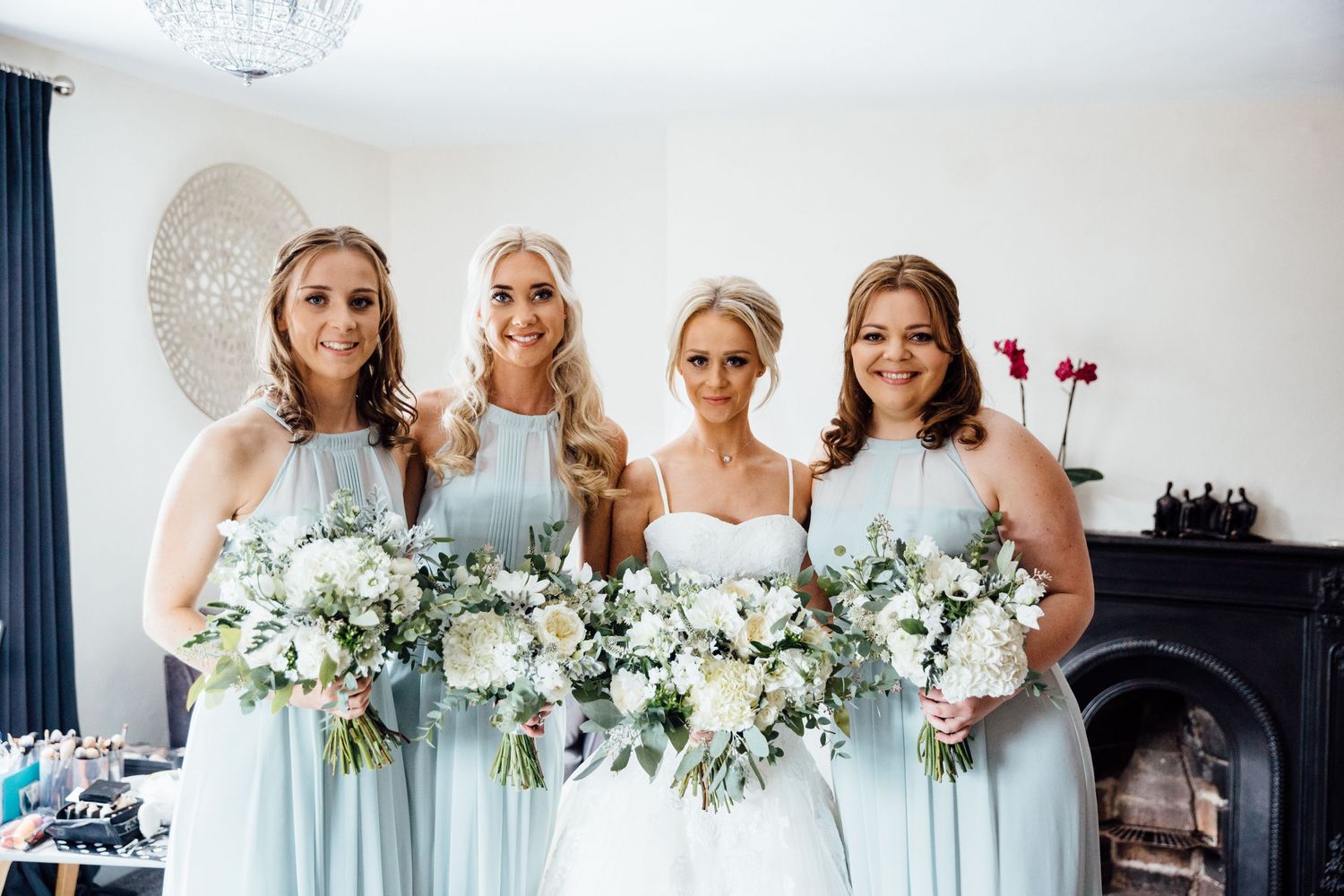 Bridesmaids in pale blue dresses hold white floral bouquets against white wall.