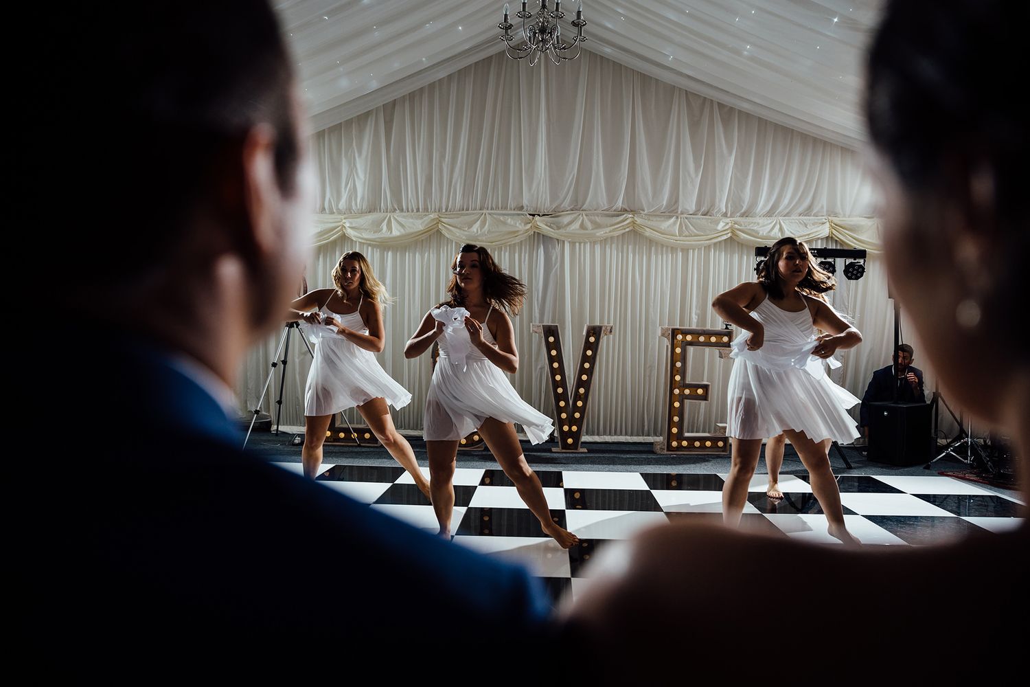 Three performers in white dresses dance on a checkered floor under a white tent.