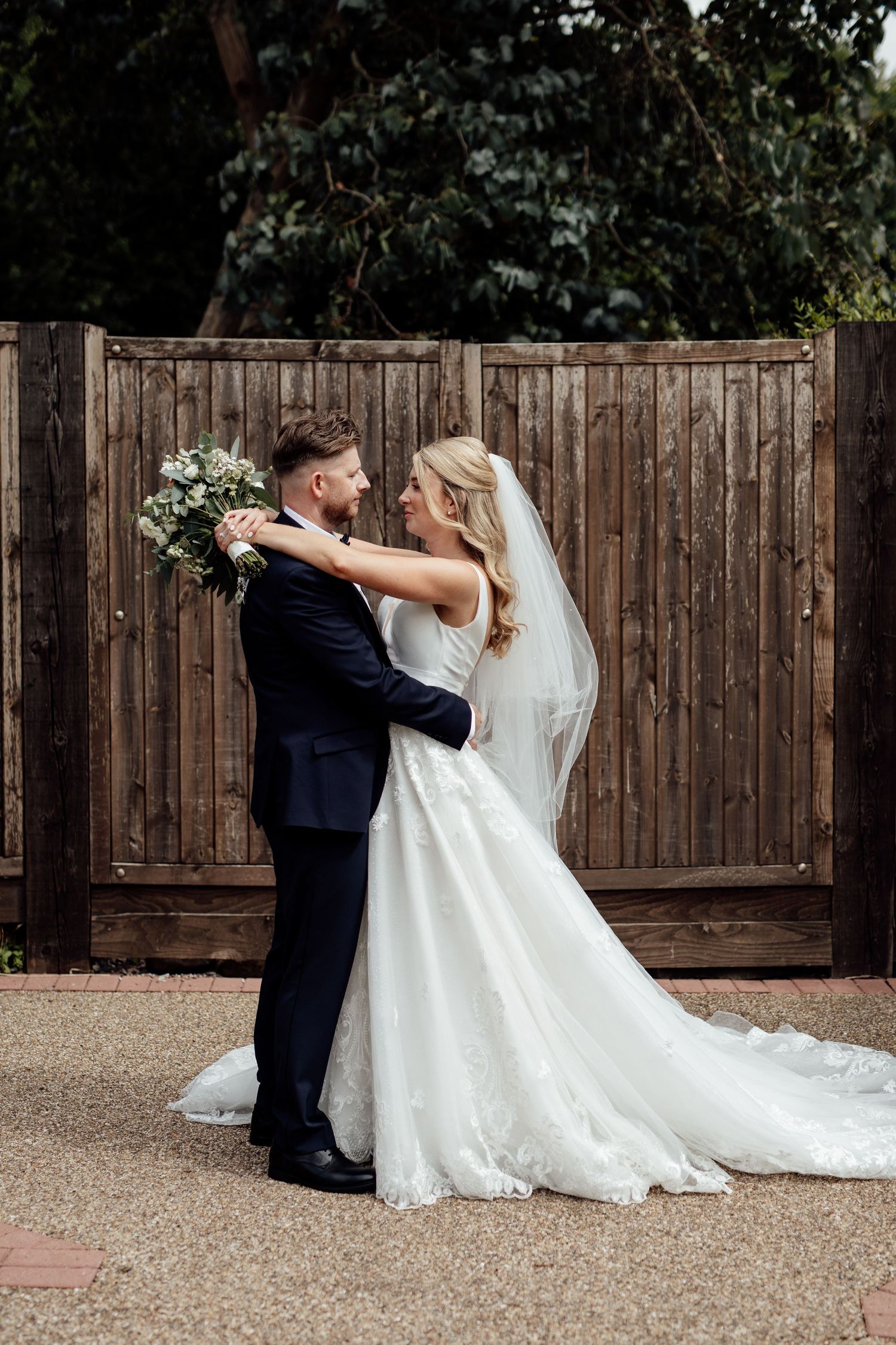 Romantic wedding portrait against wooden fence as couple embraces outdoors.