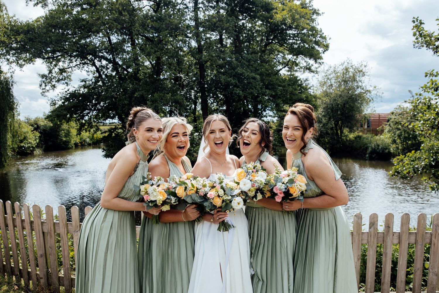Wedding party poses by a lakeside garden with white picket fence and trees in background.