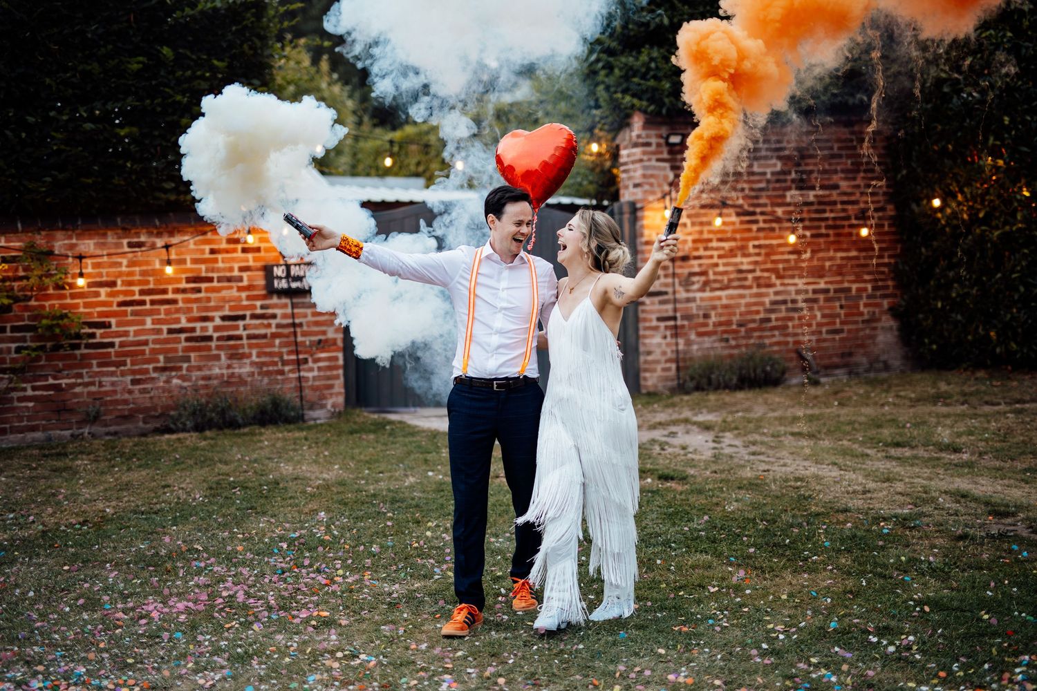 Couple celebrates with colorful smoke bombs against brick wall backdrop in garden setting.