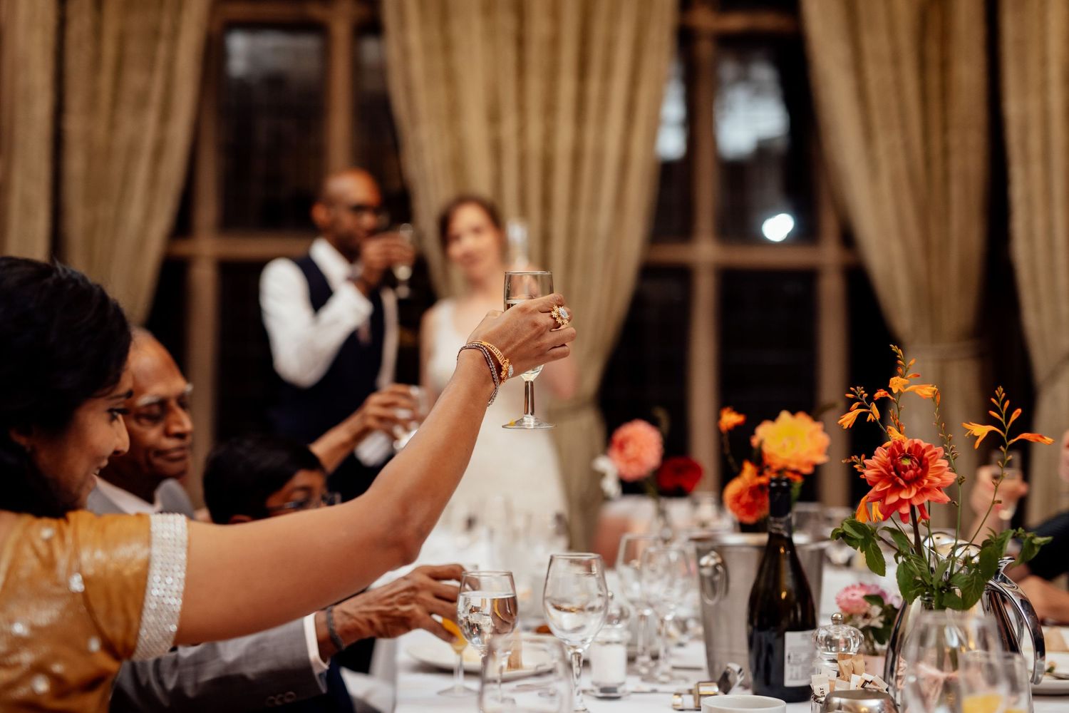 Wedding reception guests raise glasses in a toast at an elegant dinner table with floral centerpieces.