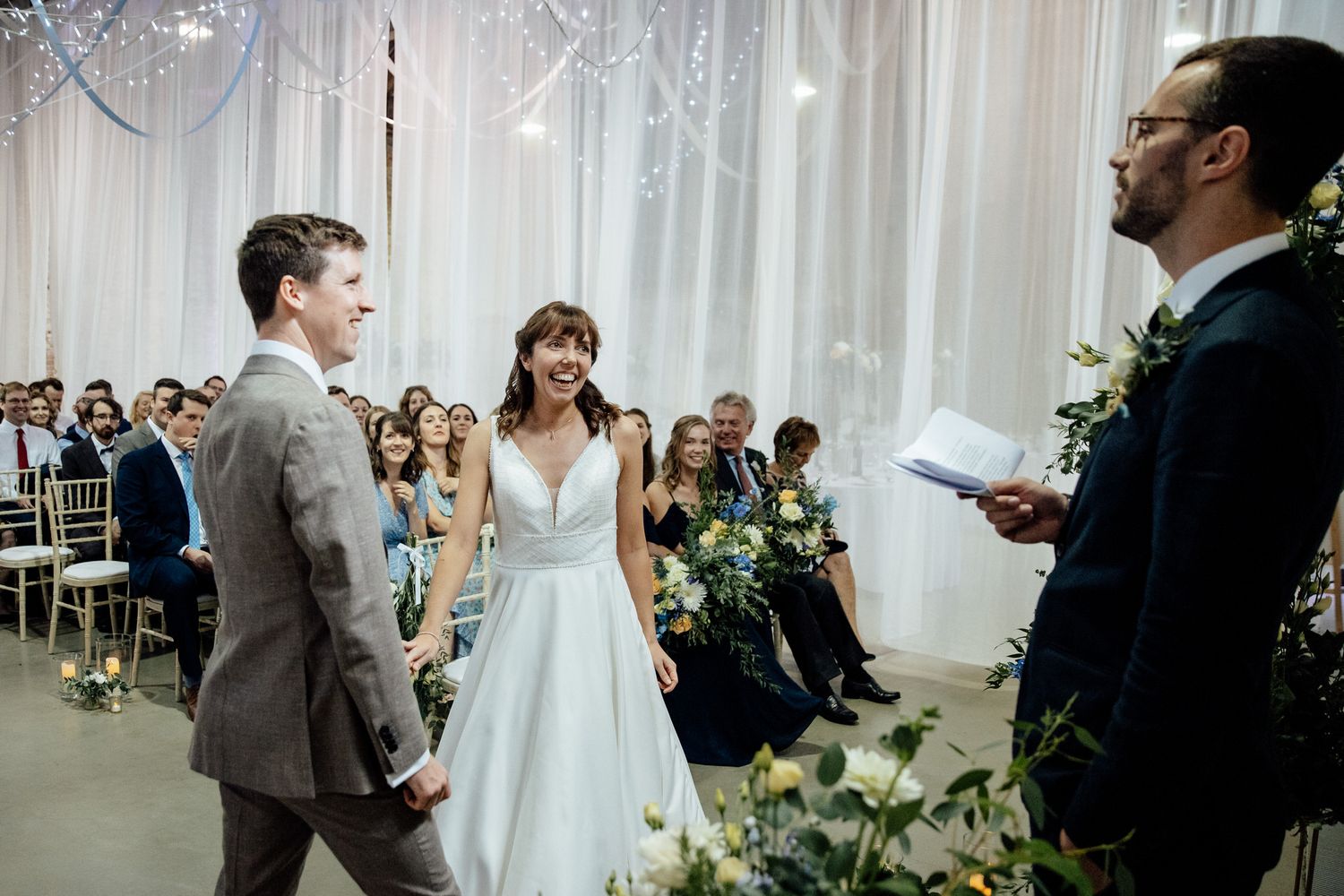 Wedding ceremony in progress inside a white-draped venue with floral decorations.