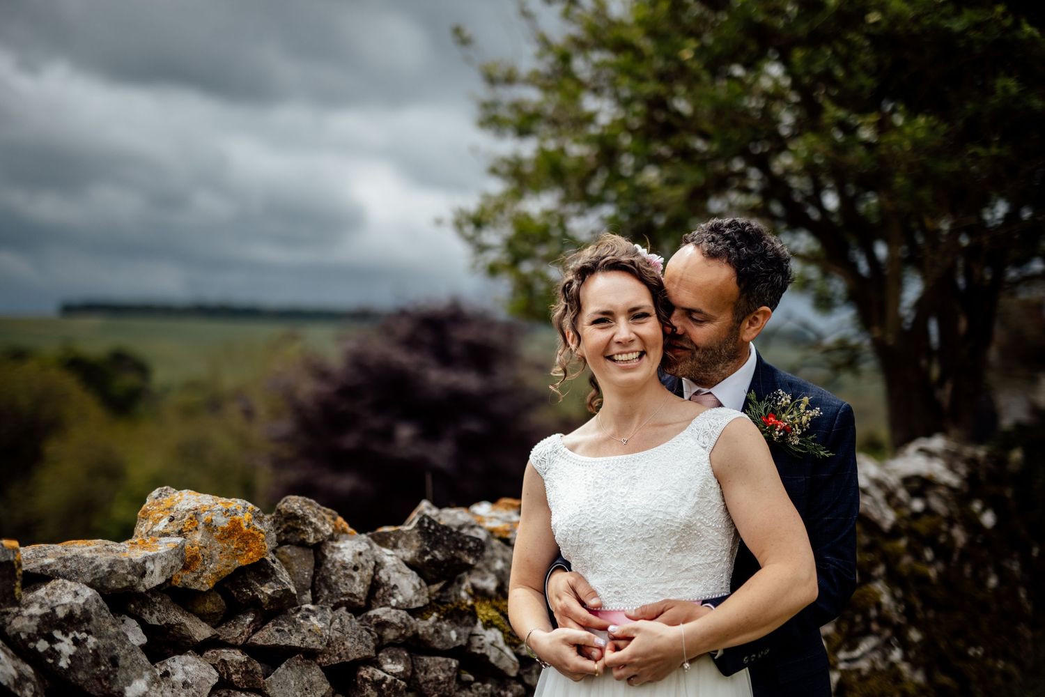 A couple shares an intimate moment against a scenic countryside backdrop with stone walls.