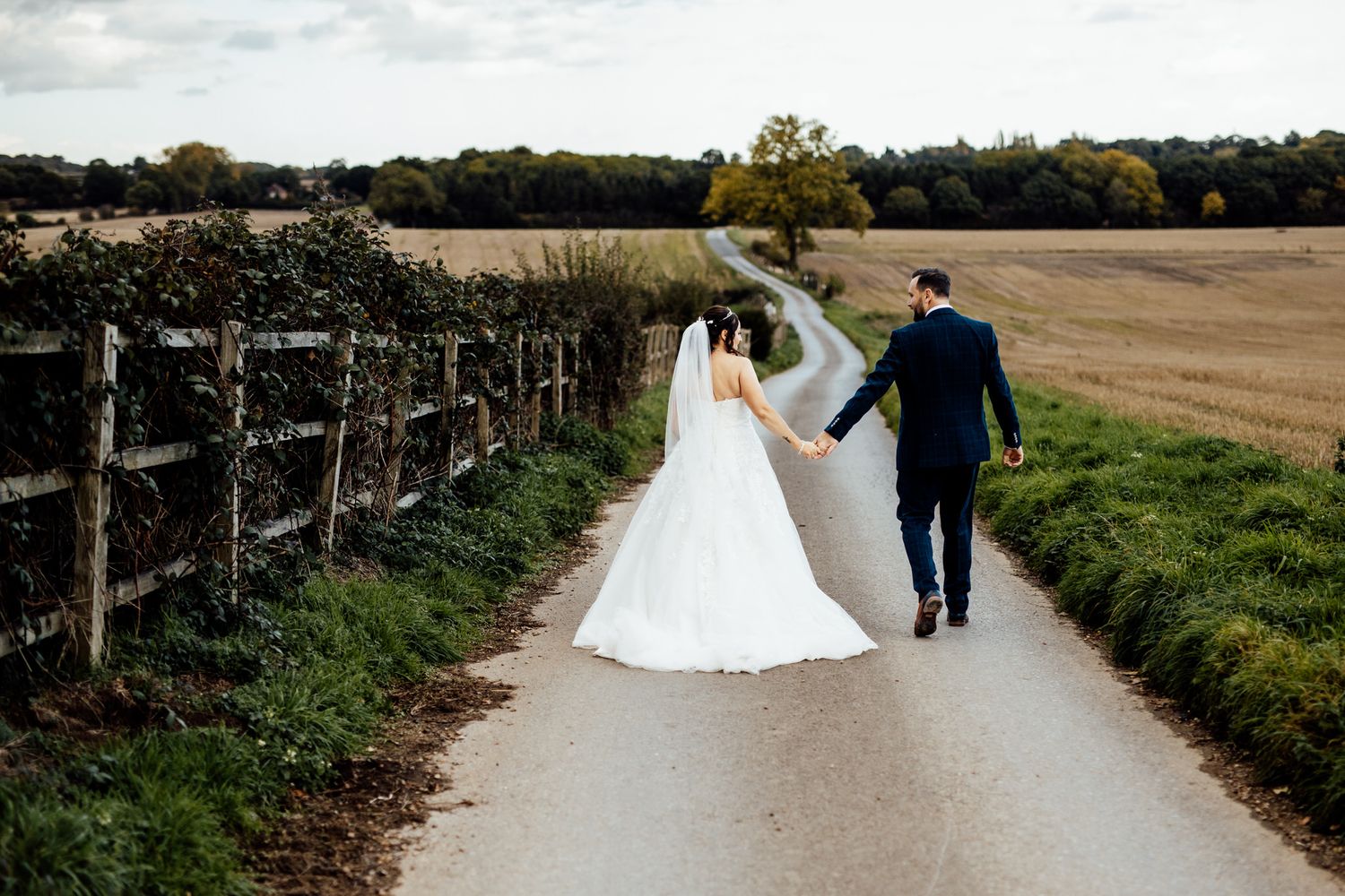 A couple walks hand in hand down a country lane bordered by fields and wooden fencing.