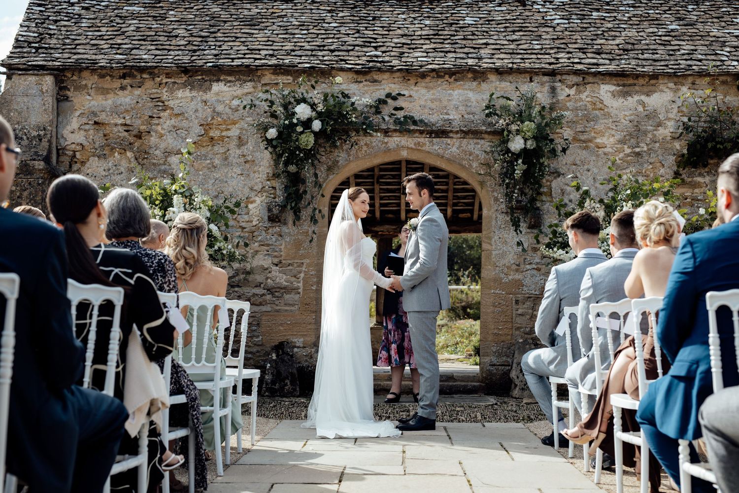 Wedding guests seated at outdoor ceremony with stone wall backdrop.