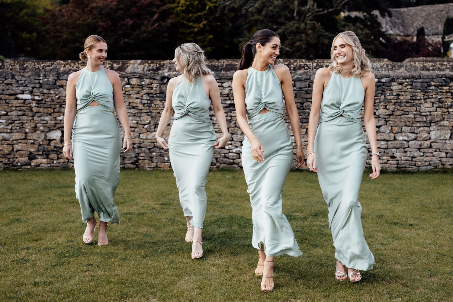 Four bridesmaids in matching mint green dresses walking together on grass near stone wall.