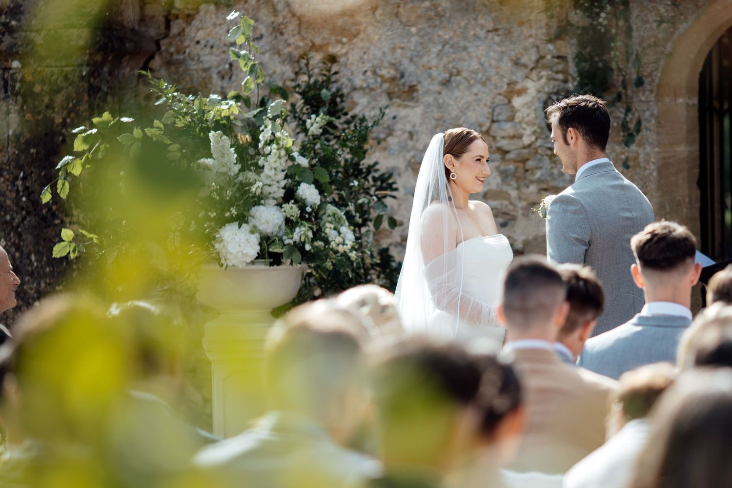 A wedding ceremony takes place outdoors with white floral arrangements against a stone wall backdrop.