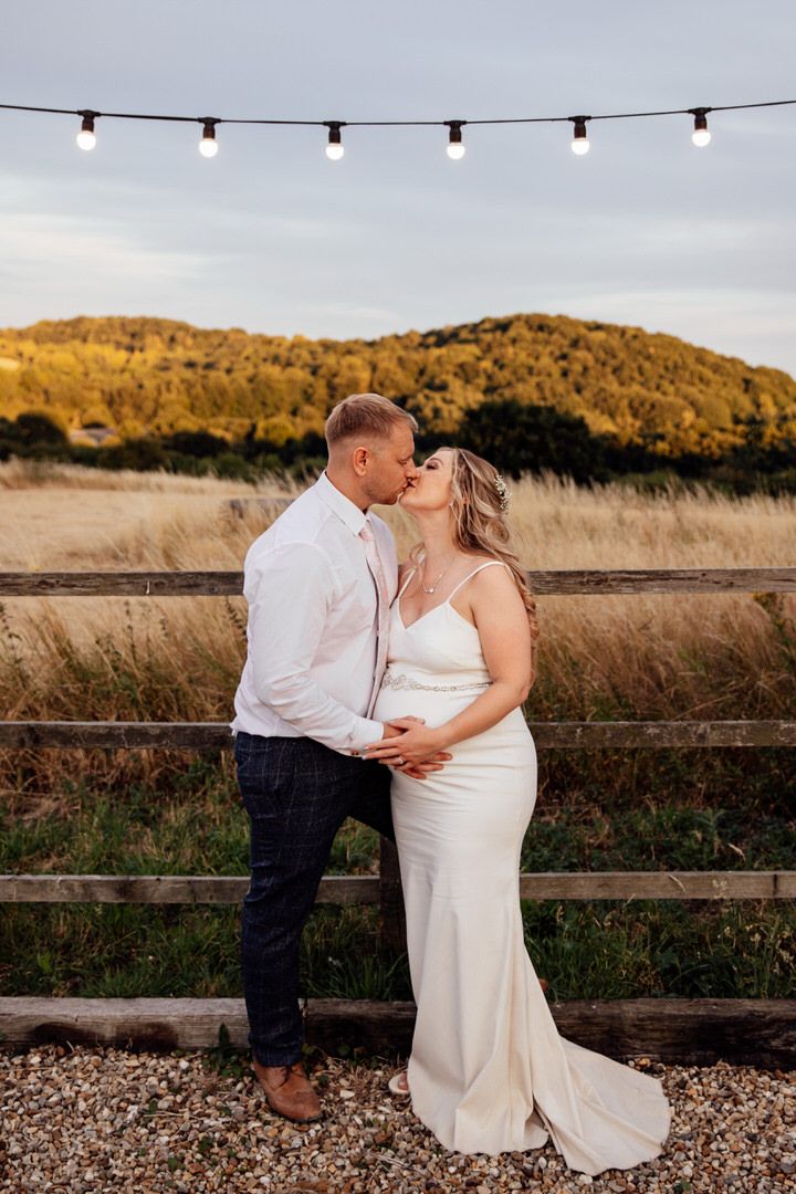 Couple sharing an intimate sunset moment against a rustic fence with string lights overhead.
