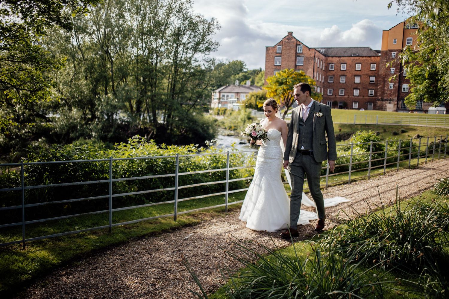 A couple poses for wedding photos on a scenic path with brick buildings in the background.