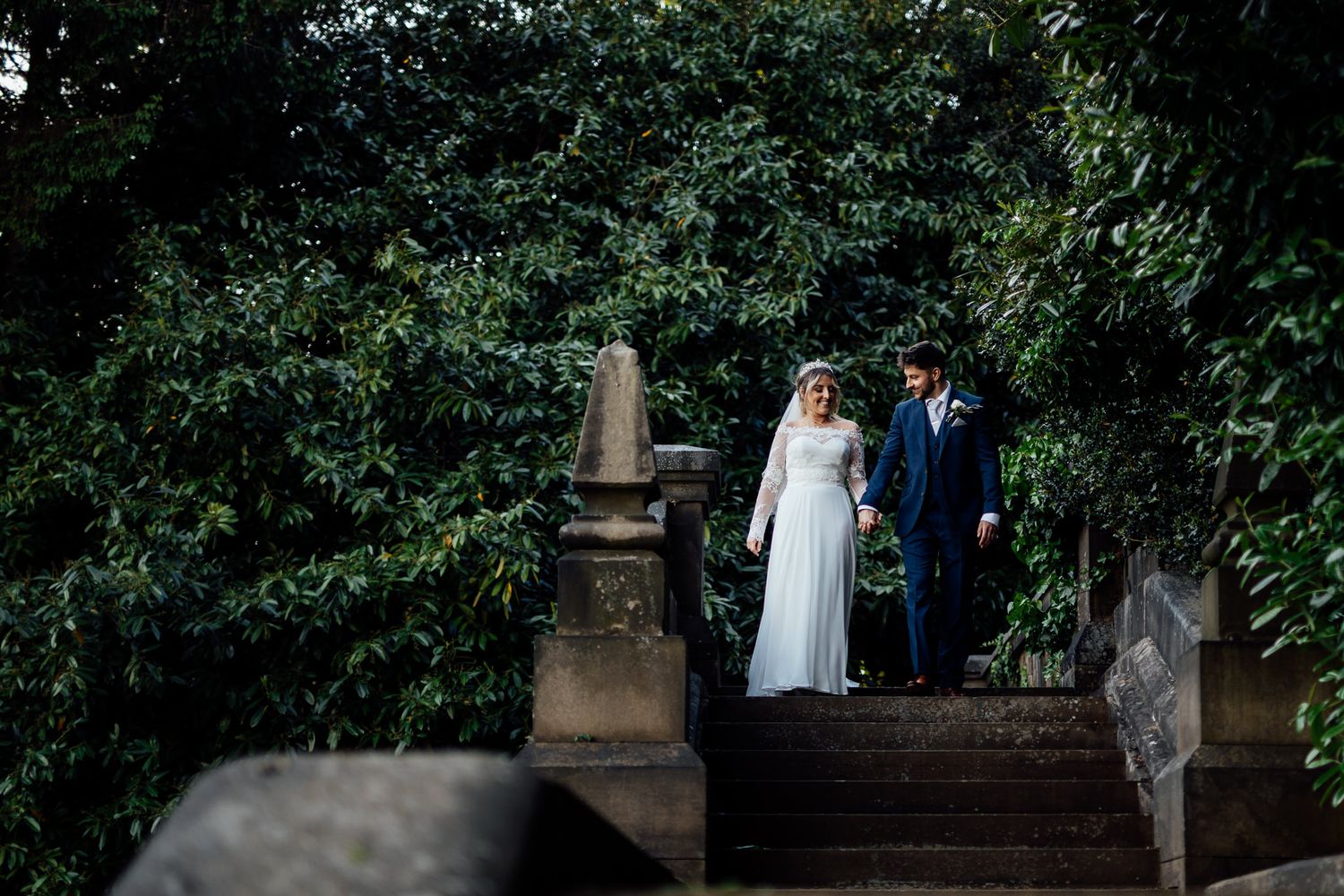Couple walking together in a garden setting with stone architecture and greenery.