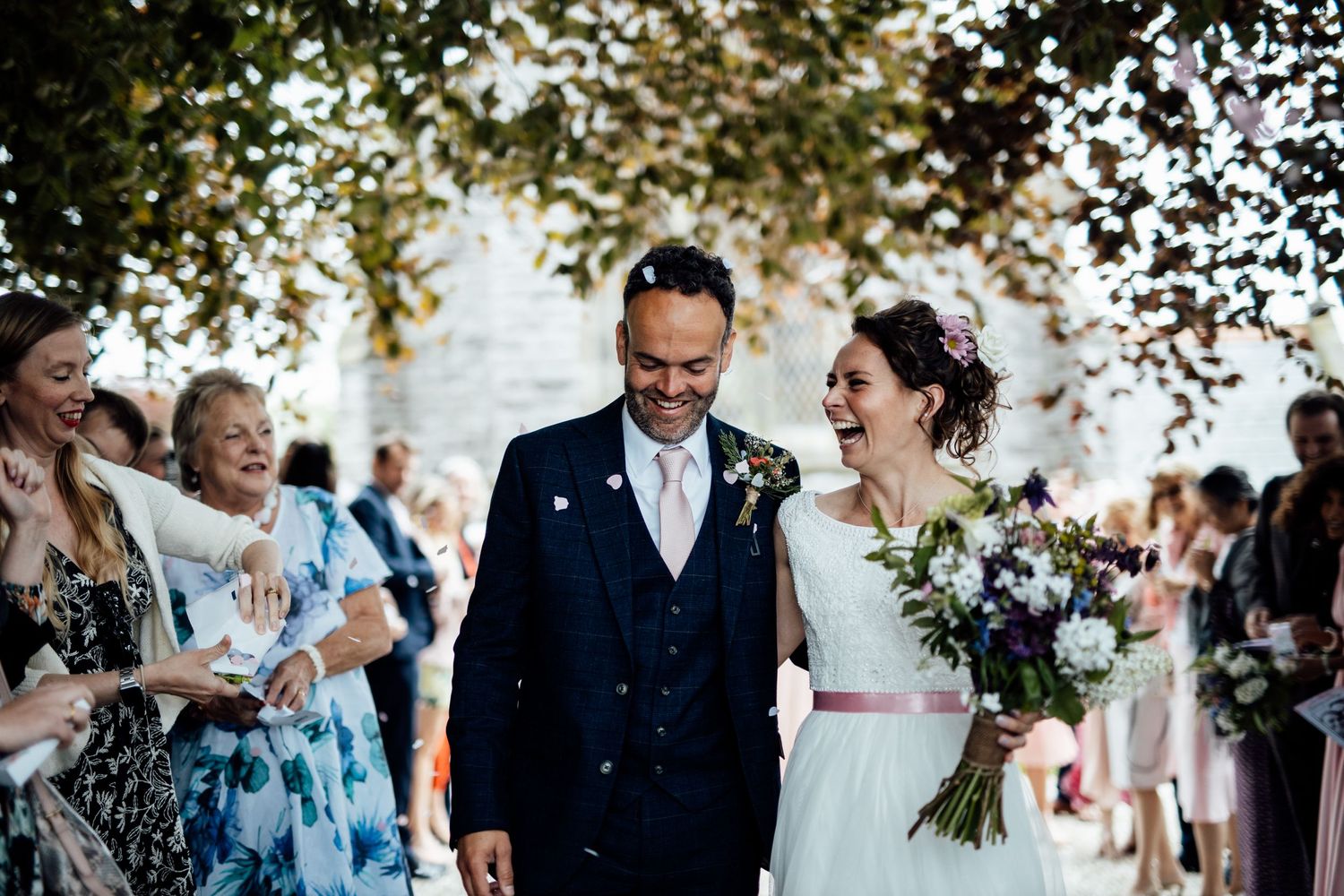 Wedding guests celebrate outdoors under tree branches on a sunny day.