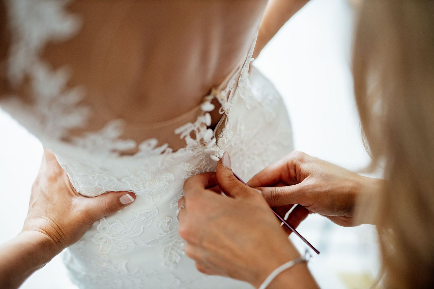 Close-up details of a white lace wedding dress being buttoned up in natural light.
