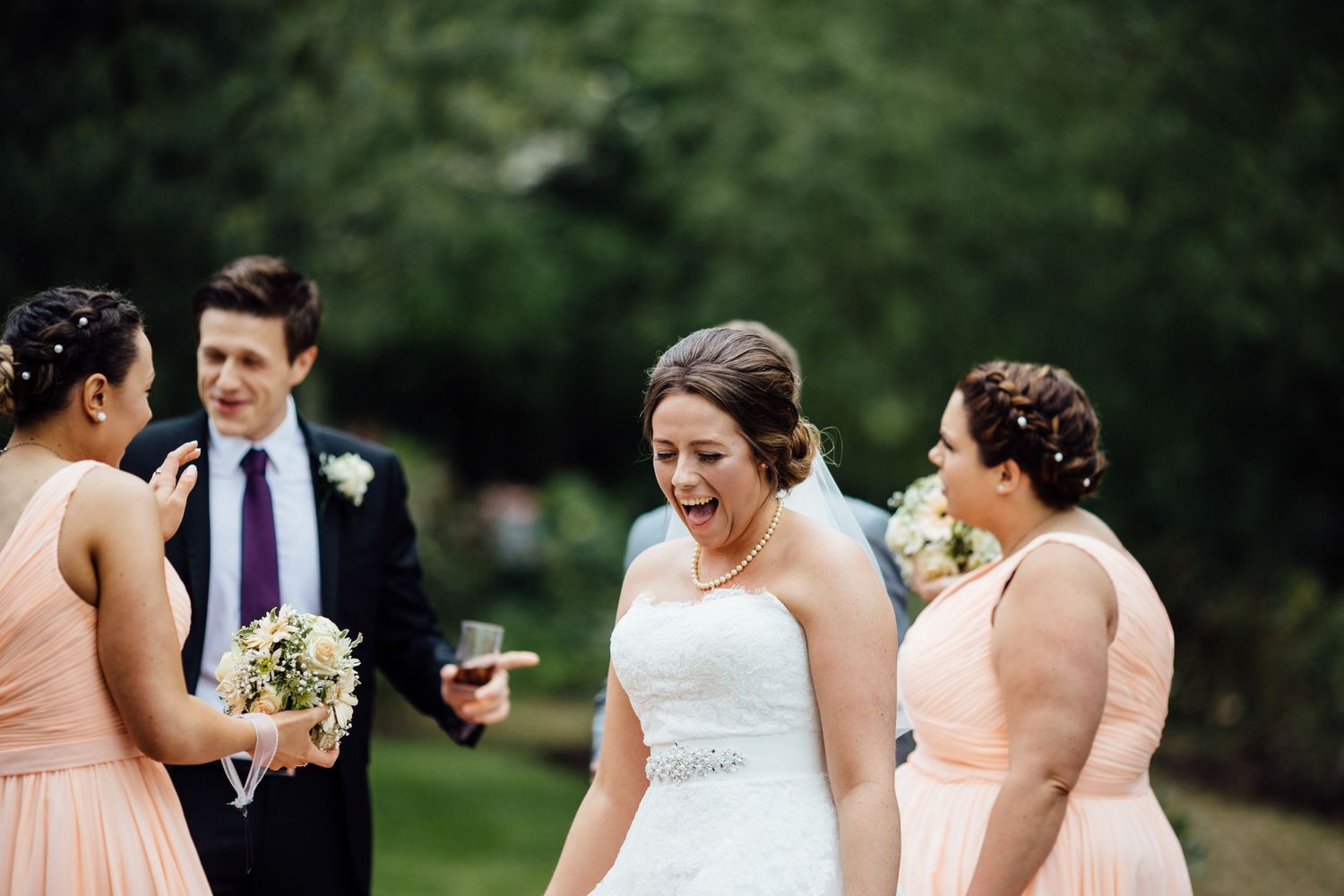 Wedding party in peach dresses and gray suits pose outdoors on green lawn.
