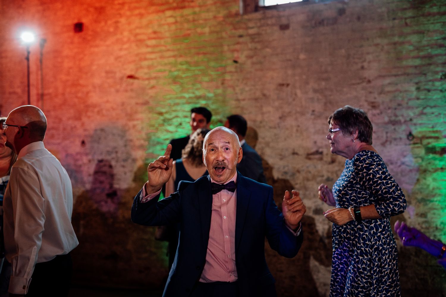 Wedding celebration in a rustic brick venue with colorful mood lighting and dancing guests.