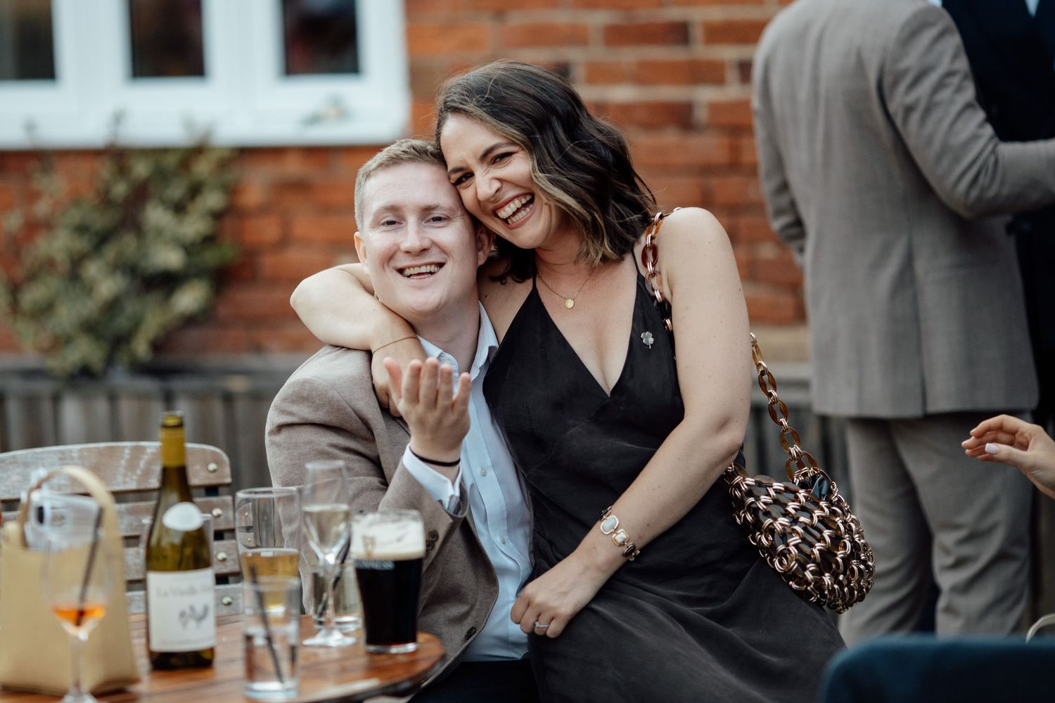 Wedding guests enjoying drinks and conversation at an outdoor reception with brick wall backdrop.