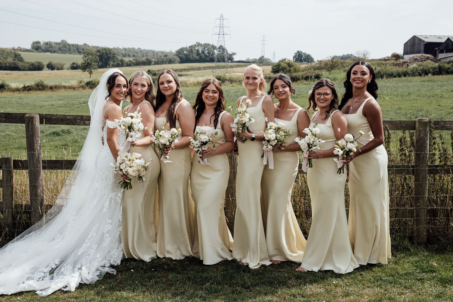 Bridesmaids in cream-colored dresses pose together in a rural outdoor setting.