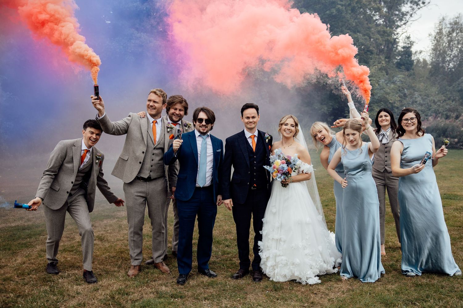 Wedding party poses with colorful smoke bombs in an outdoor setting.