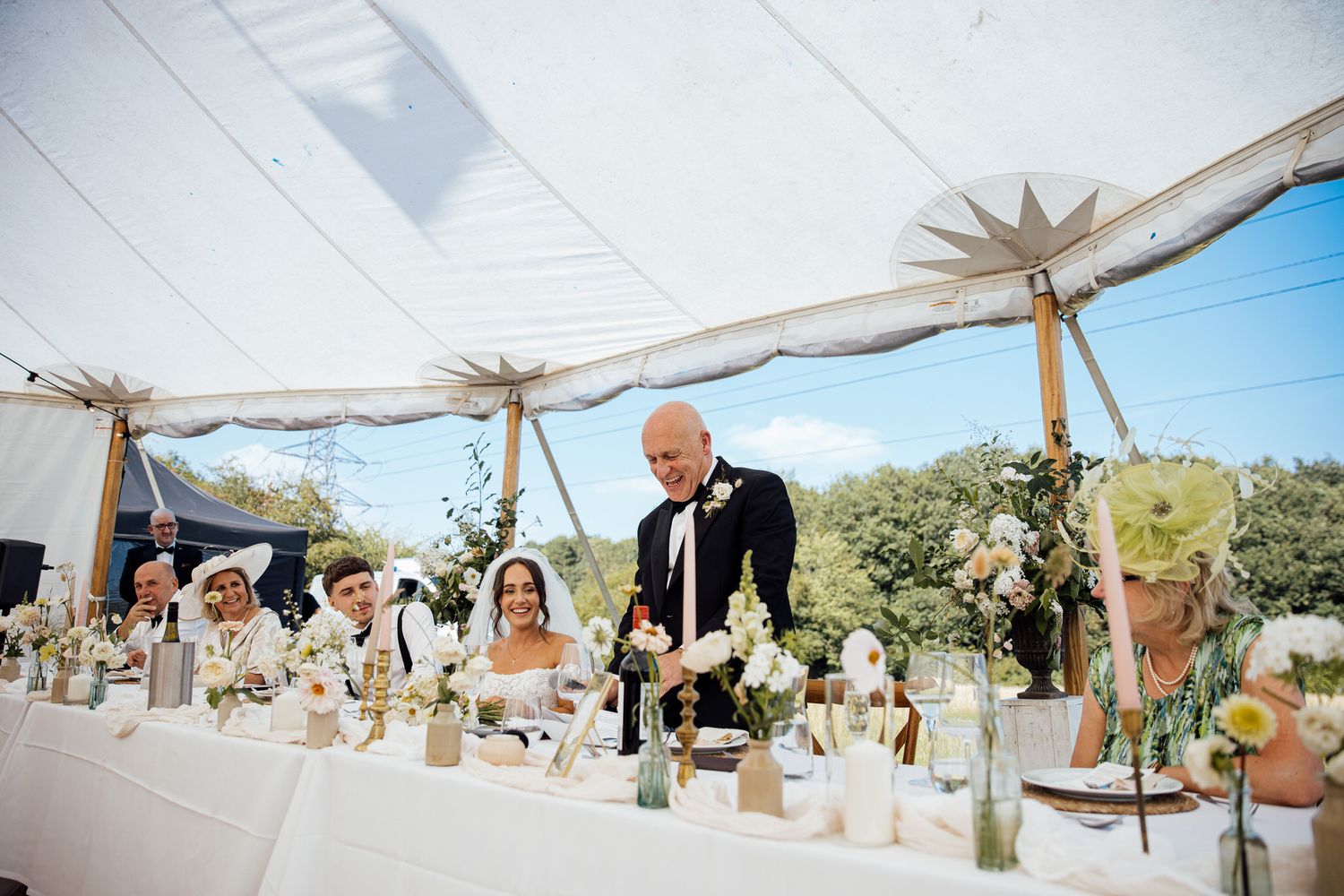 Wedding couple and guests seated at long table under tent with rustic decor and centerpieces.