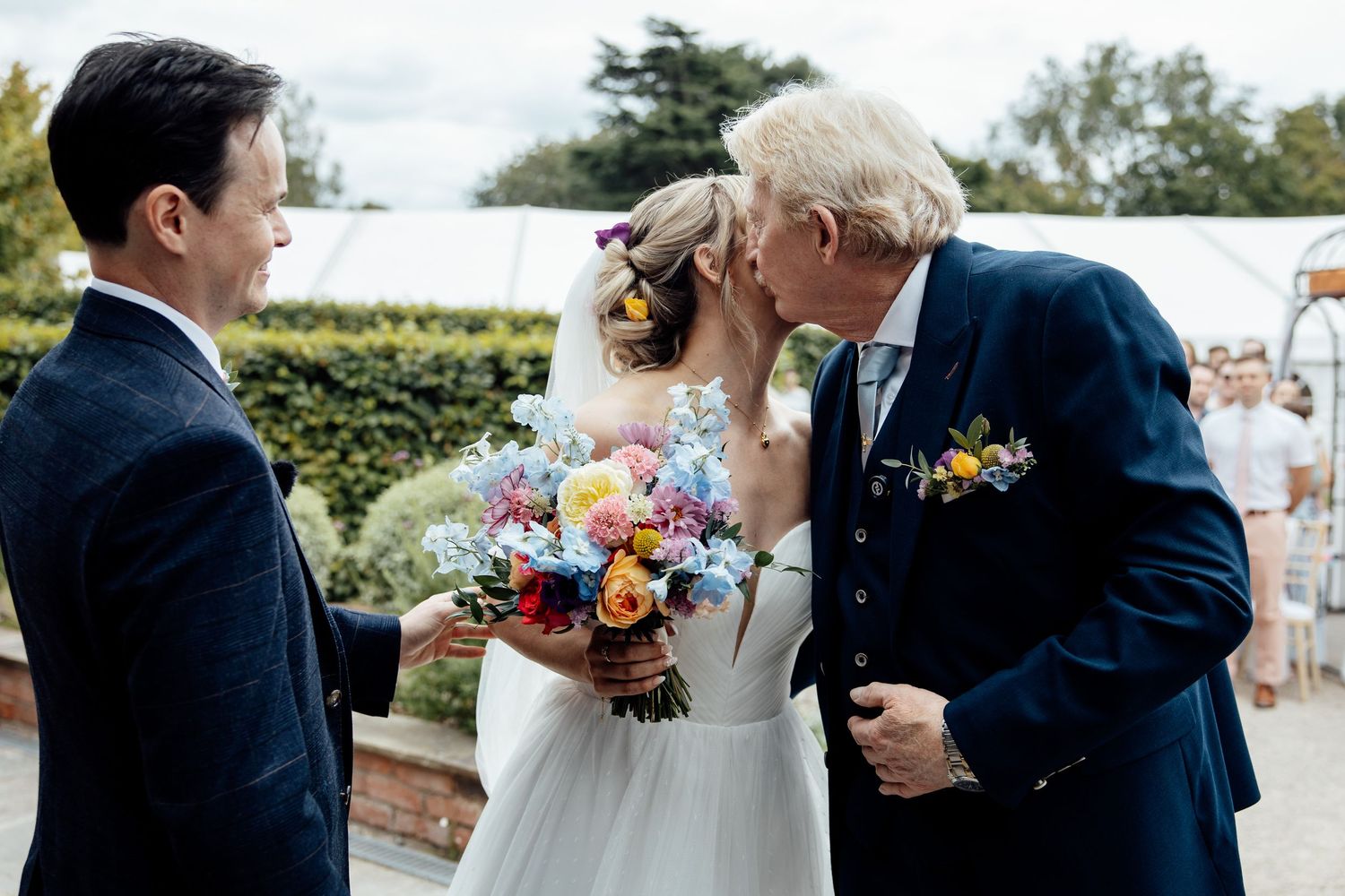 Intimate wedding ceremony moment with couple and officiant surrounded by floral bouquets outdoors.
