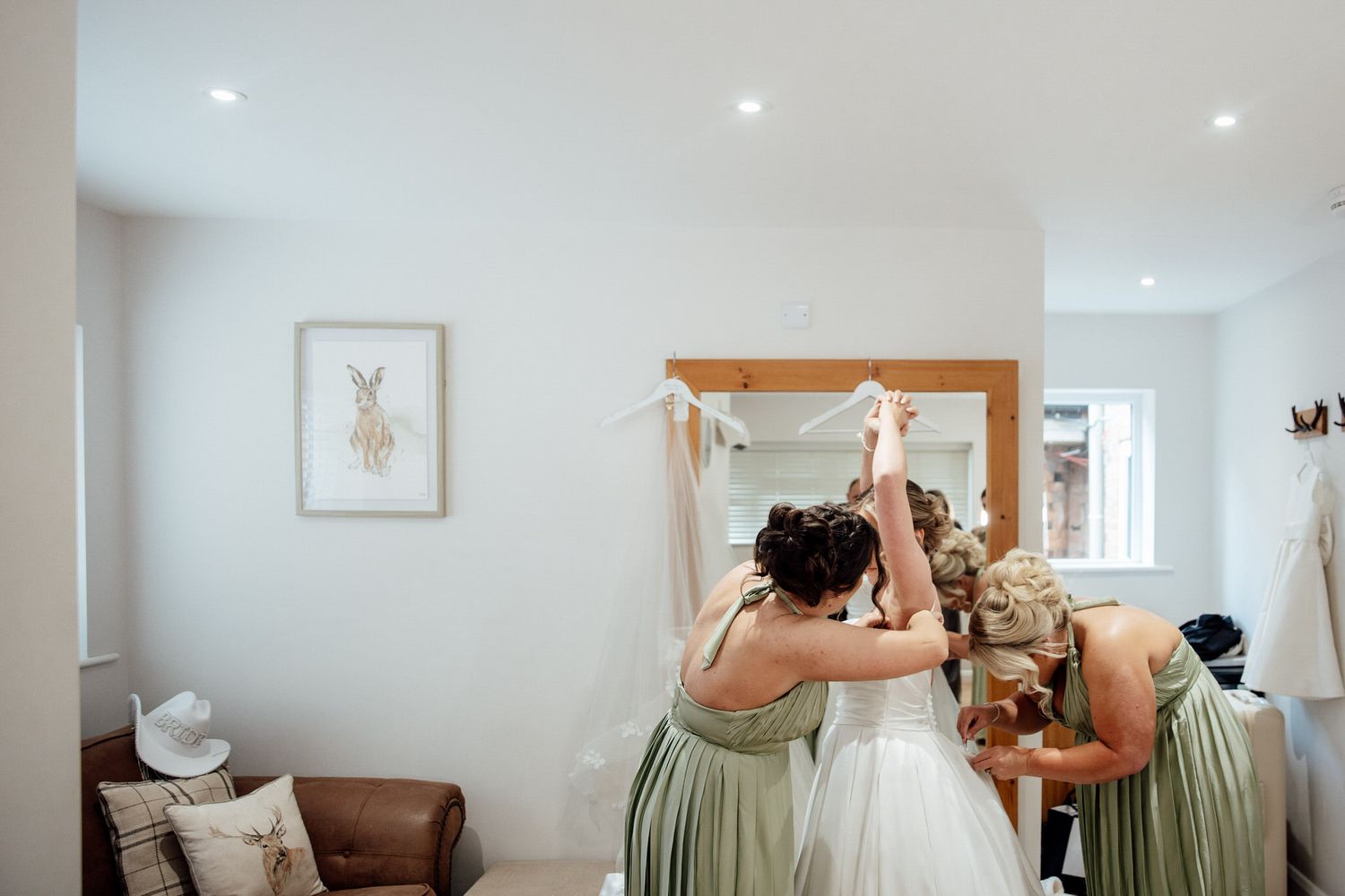 Bridesmaids in sage green dresses help the bride prepare for her wedding day in a bright room.