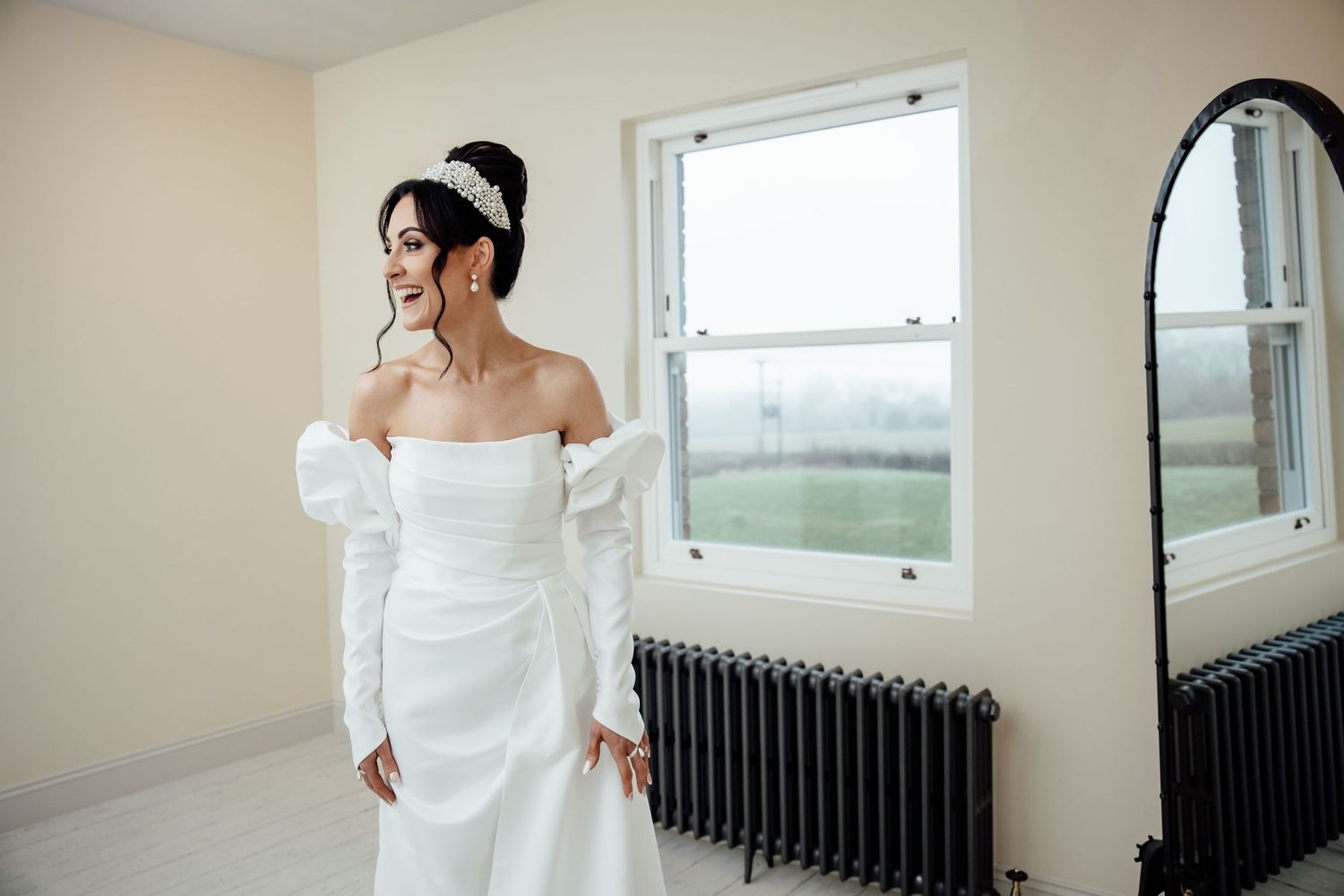Elegant off-shoulder white wedding gown shown in a bright room with vintage radiator and windows.