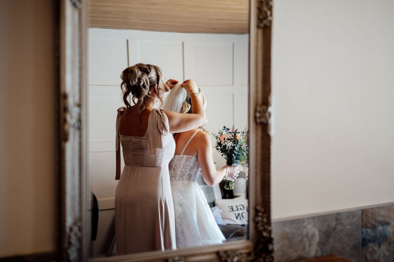 Bridesmaid helping bride with wedding dress in elegant gold-framed mirror reflection during bridal preparations.
