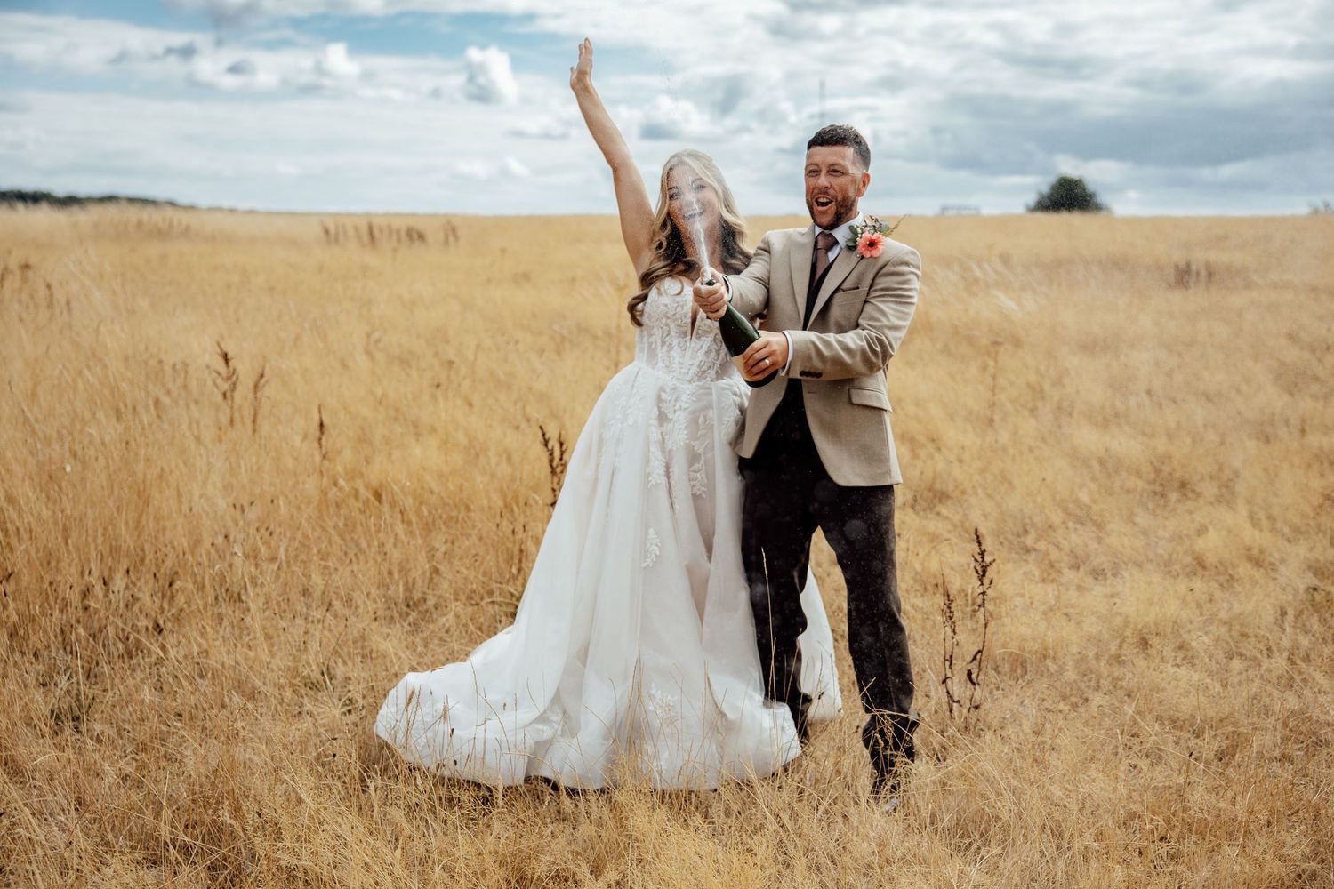 Excited newlyweds celebrate with champagne in golden wheat field under dramatic cloudy sky.