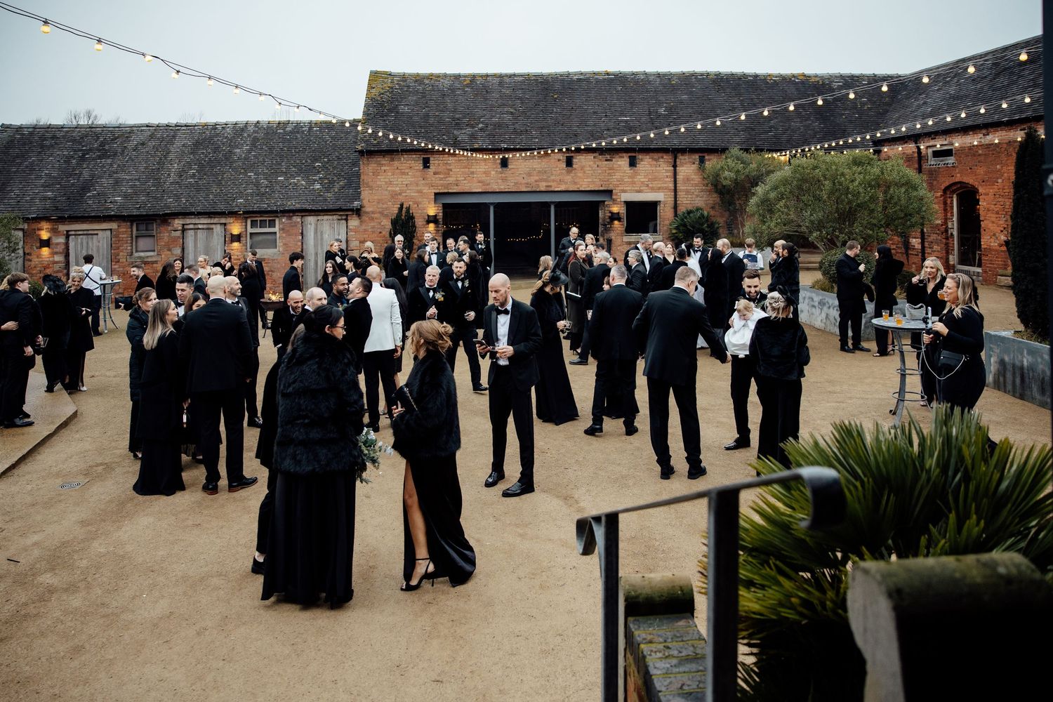 Wedding guests mingle outdoors at a rustic venue with string lights and brick buildings in the background.
