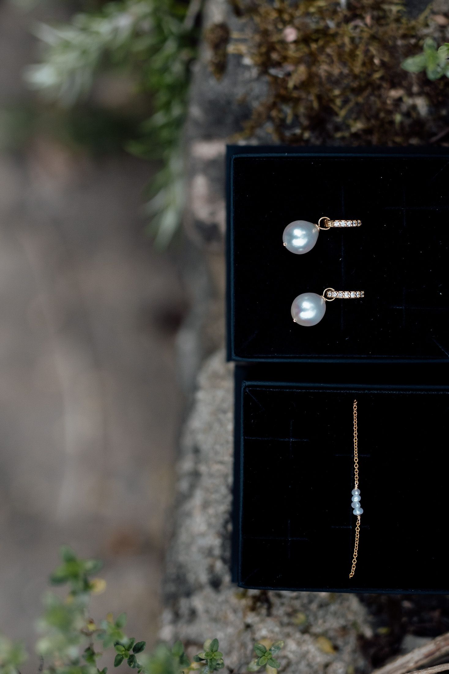 Pearl drop earrings and delicate jewelry displayed in a black presentation box against natural foliage.