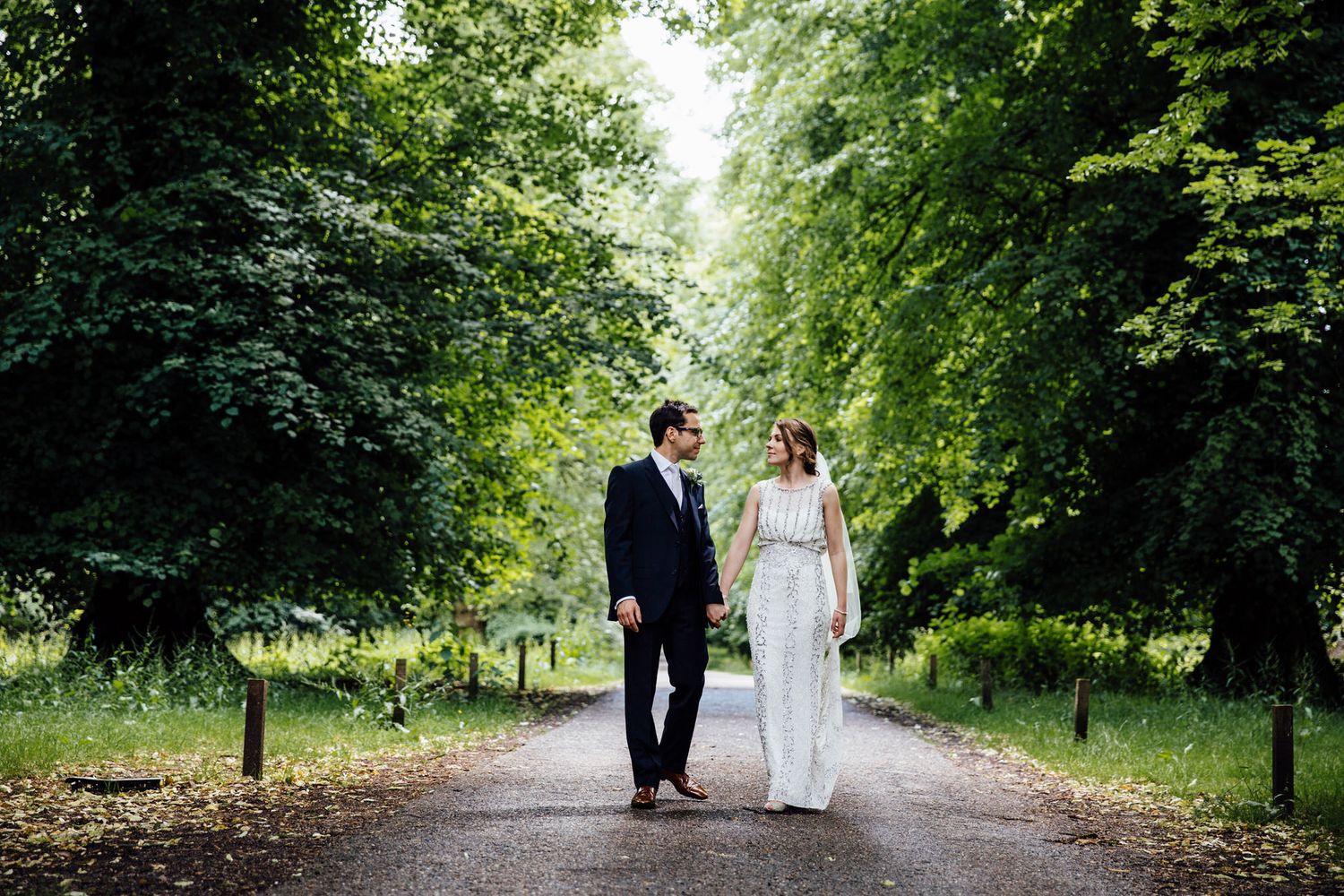 Couple walking down tree-lined path in wedding attire.
