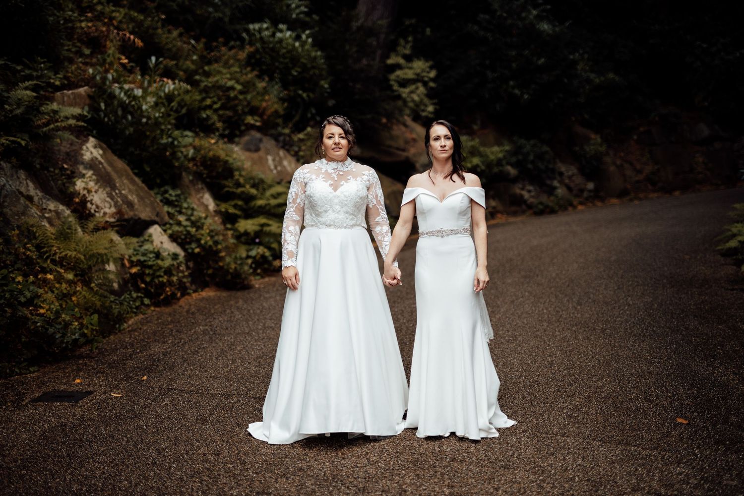 Two people in white wedding dresses walking along a dark wooded path.