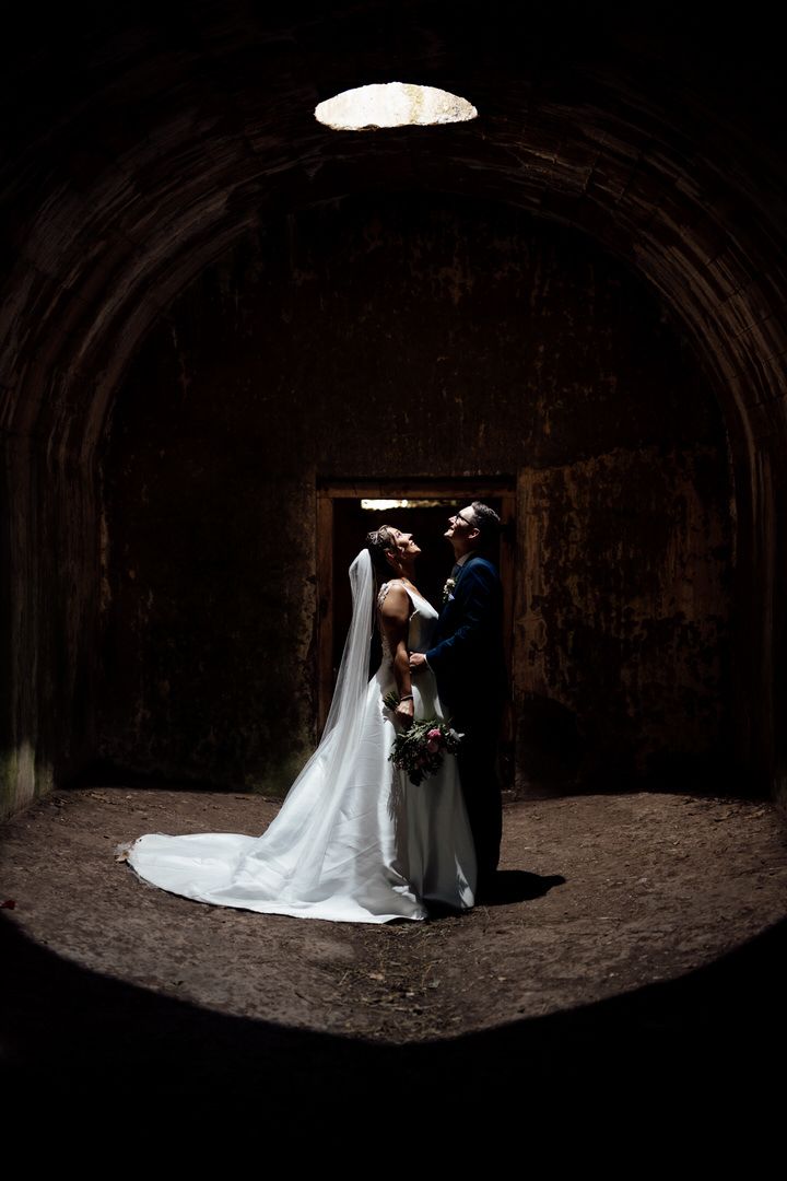 A couple in wedding attire embraces in a dramatic cave setting with natural light streaming from above.