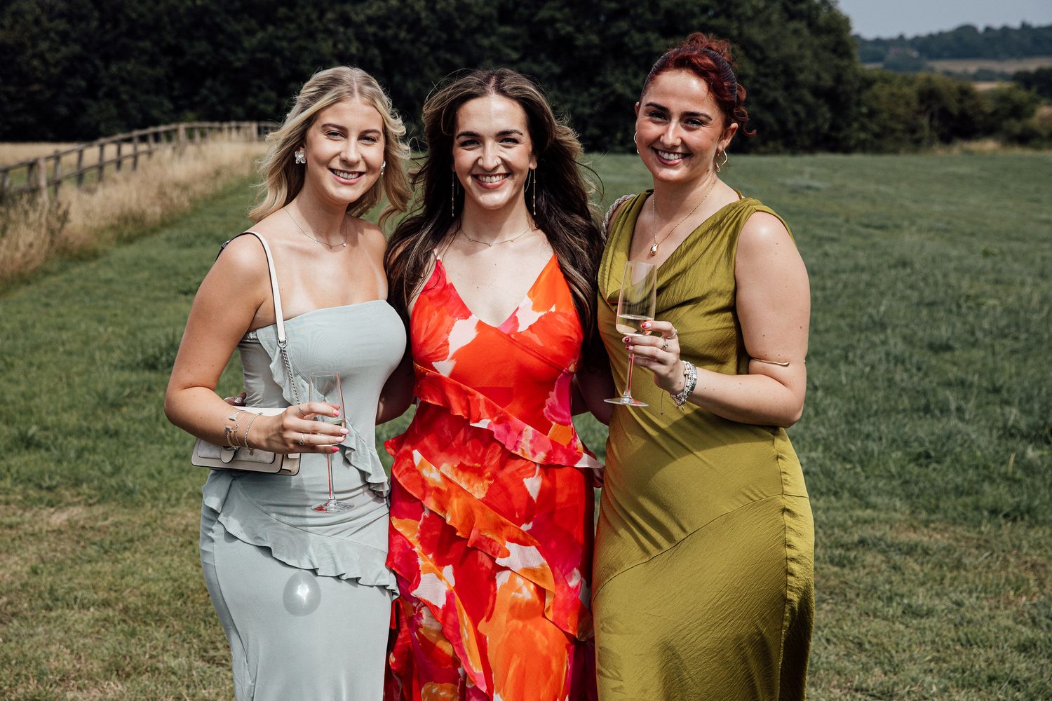 Wedding party in colorful dresses poses together on grassy field with trees in background.