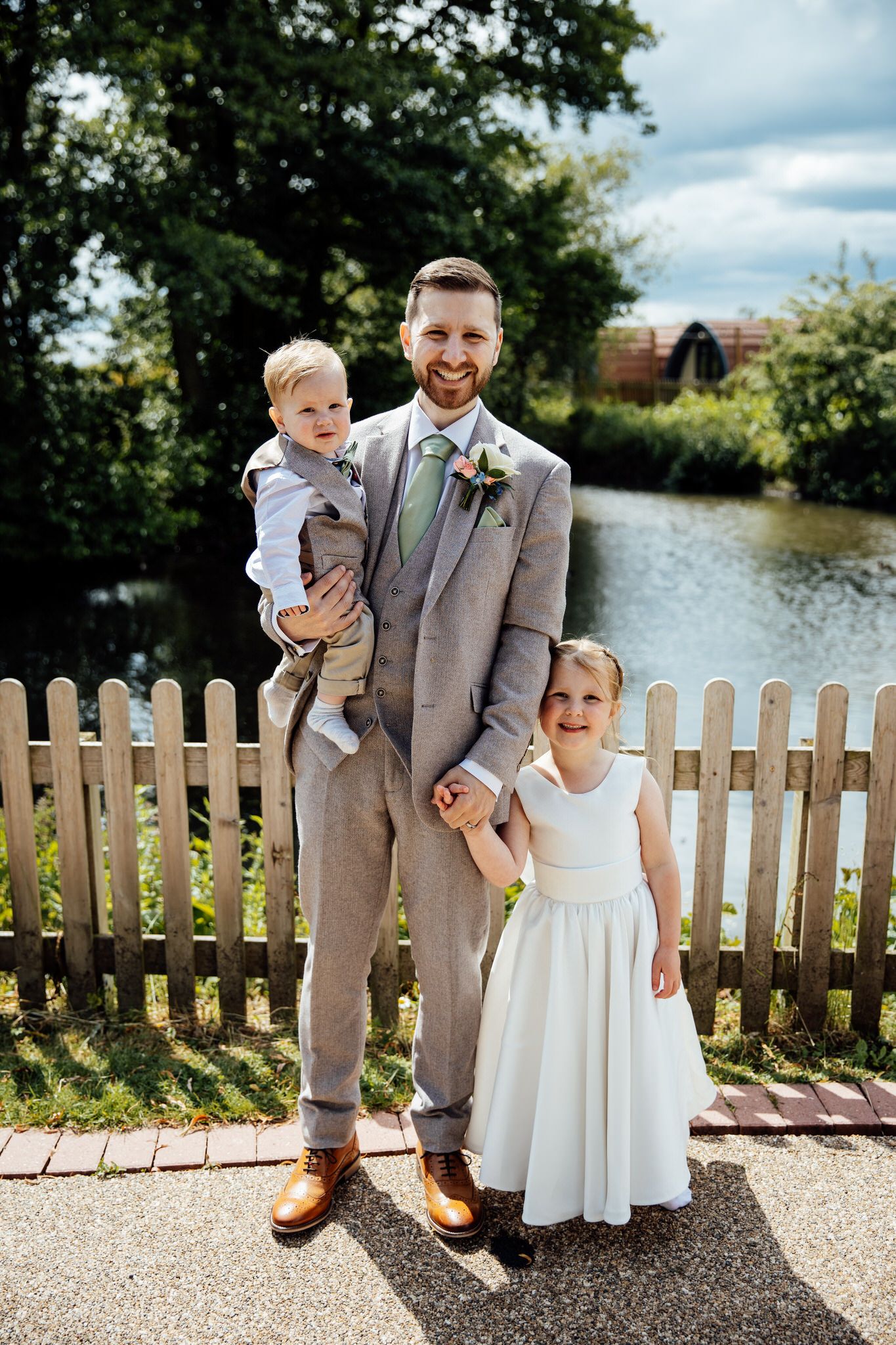 Wedding party members in formal attire posing by a white picket fence near a lake.
