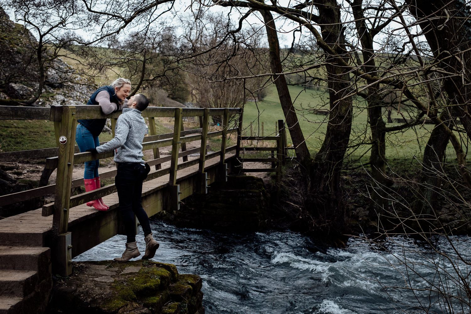 People walk across a wooden bridge over rushing water surrounded by trees.