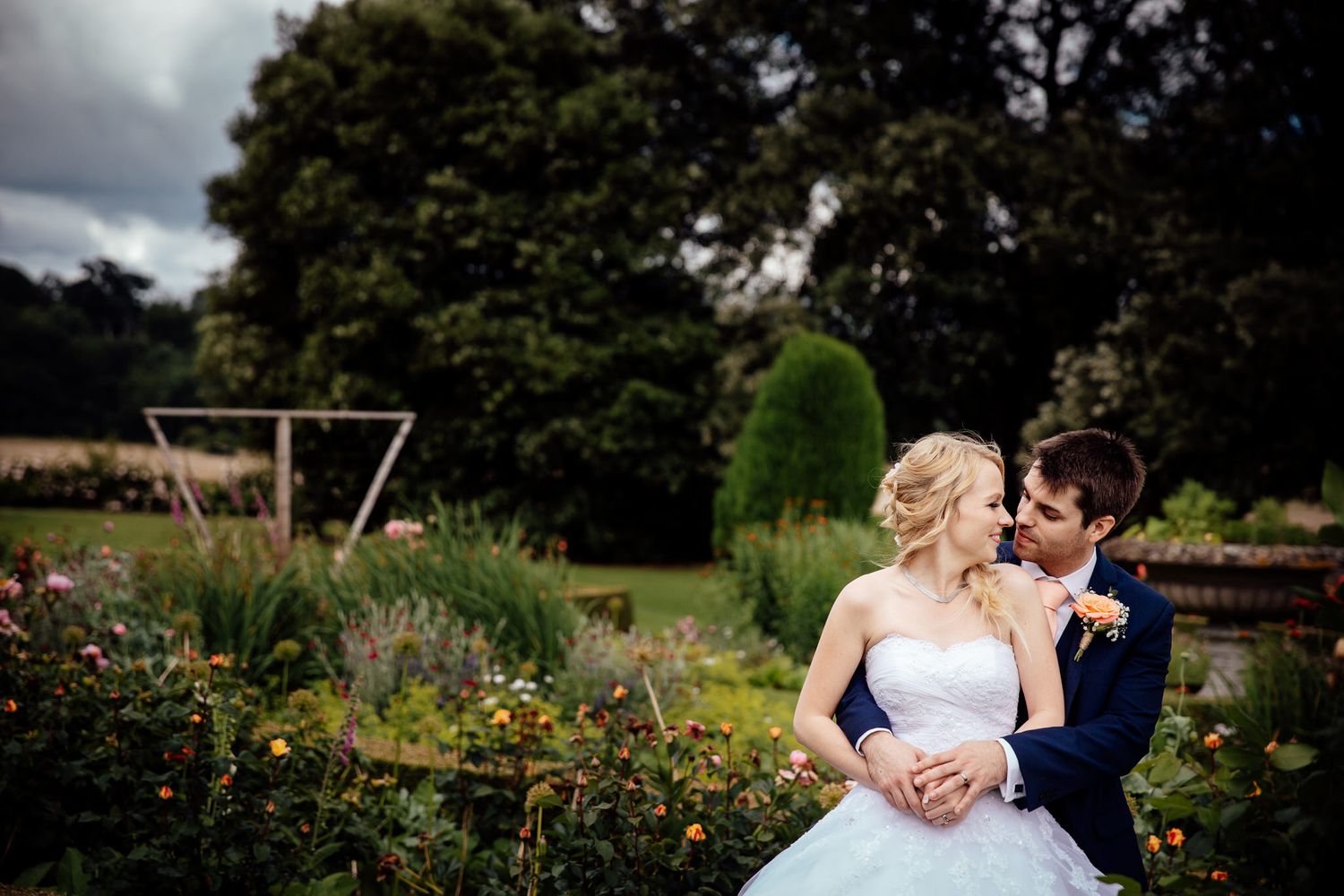 Romantic wedding couple embraces in lush garden setting under cloudy sky.