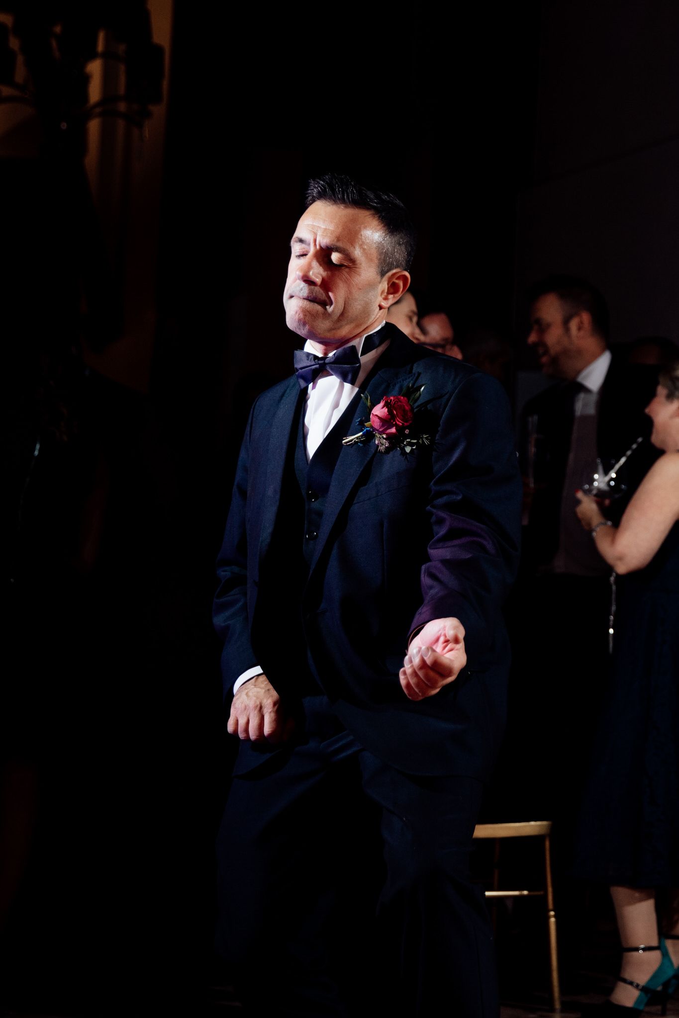 Wedding reception guests in formal attire dancing in dimly lit venue.