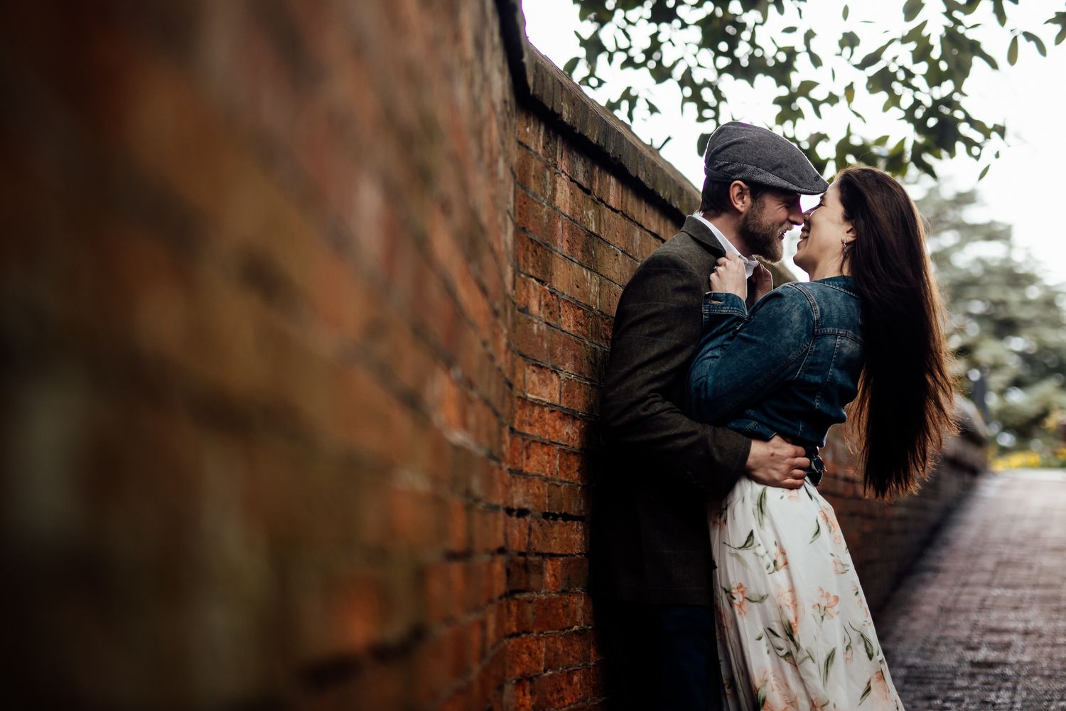 A romantic couple sits close together on a park bench surrounded by brick walls and autumn foliage.