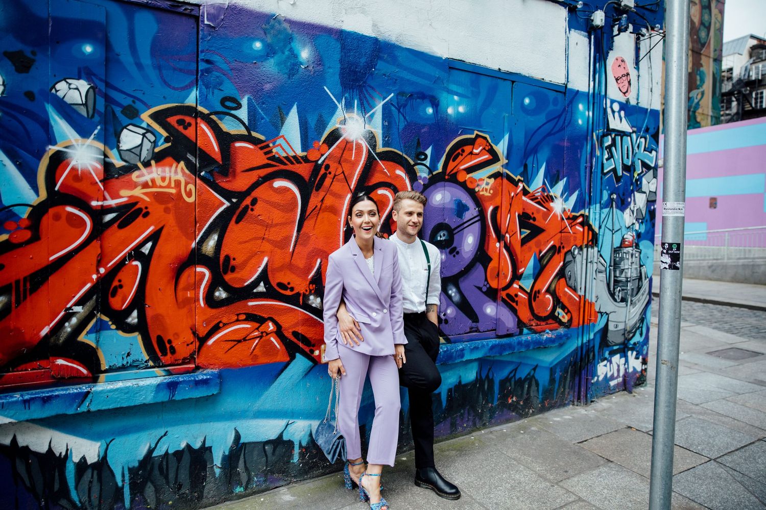 Couple posing against vibrant blue and red graffiti mural on city street.