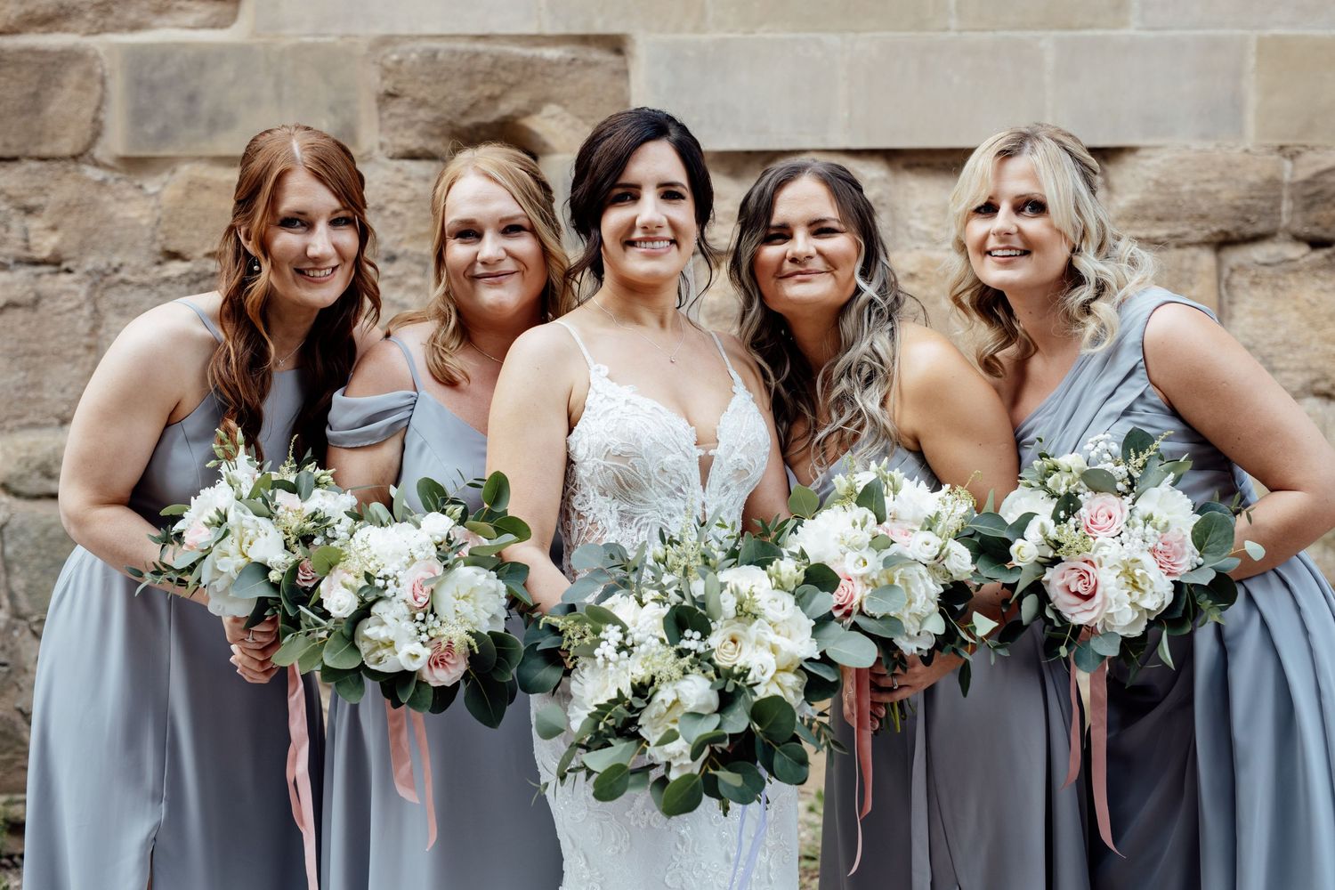 Bridesmaids in gray dresses holding white floral bouquets against stone wall.
