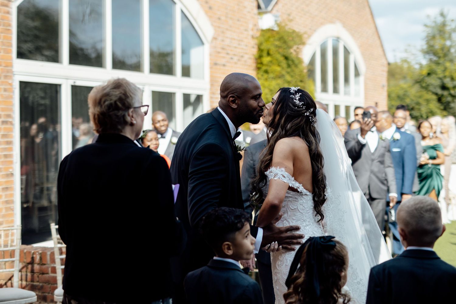 Wedding guests mingling outside a brick building during daytime celebration.