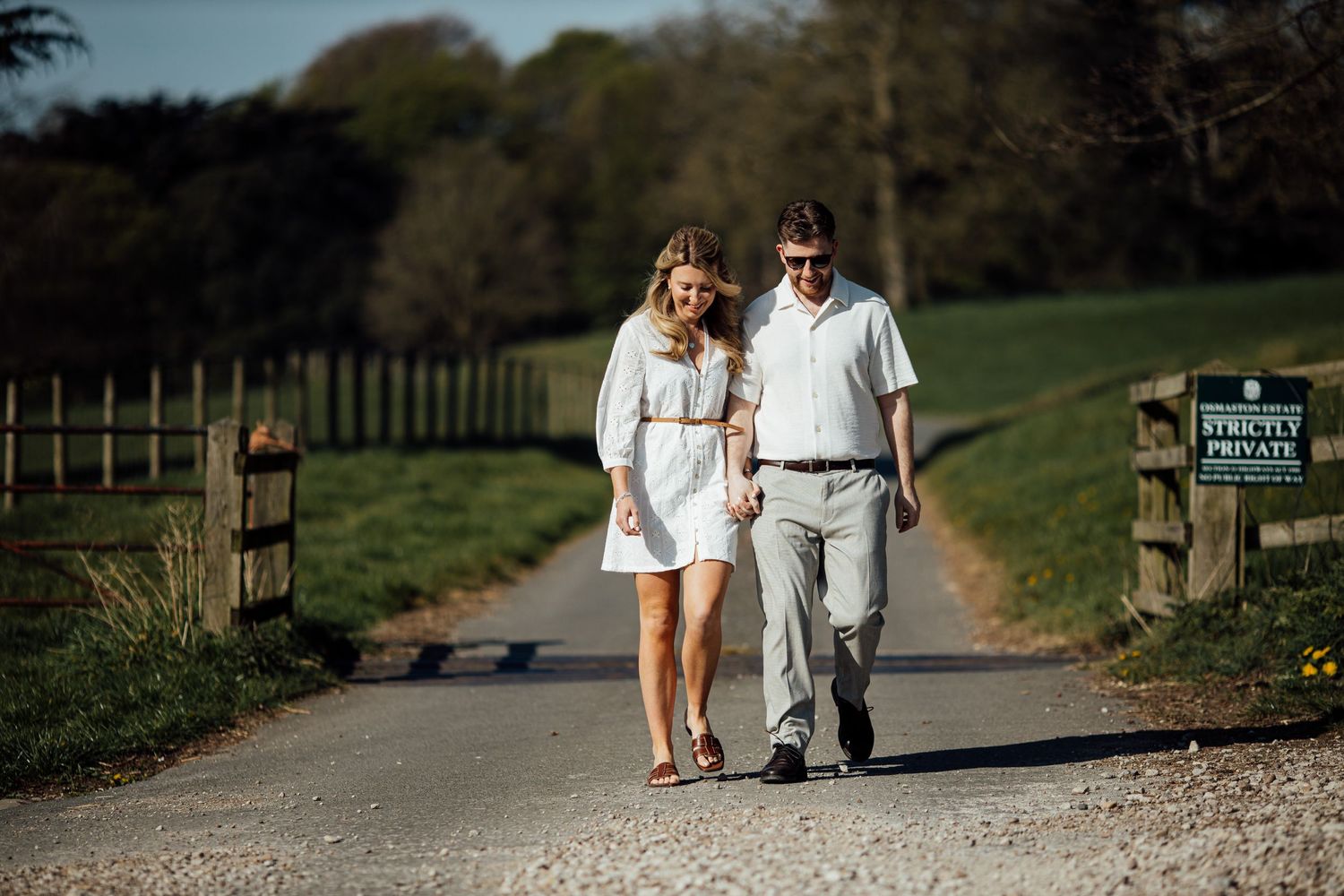 Two people strolling hand in hand along a gravel pathway surrounded by green countryside.