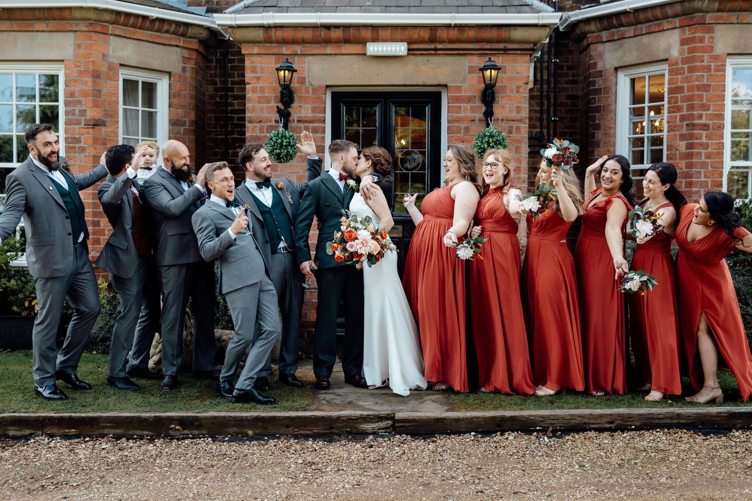 Large wedding party poses against brick building wearing red and gray formal attire.