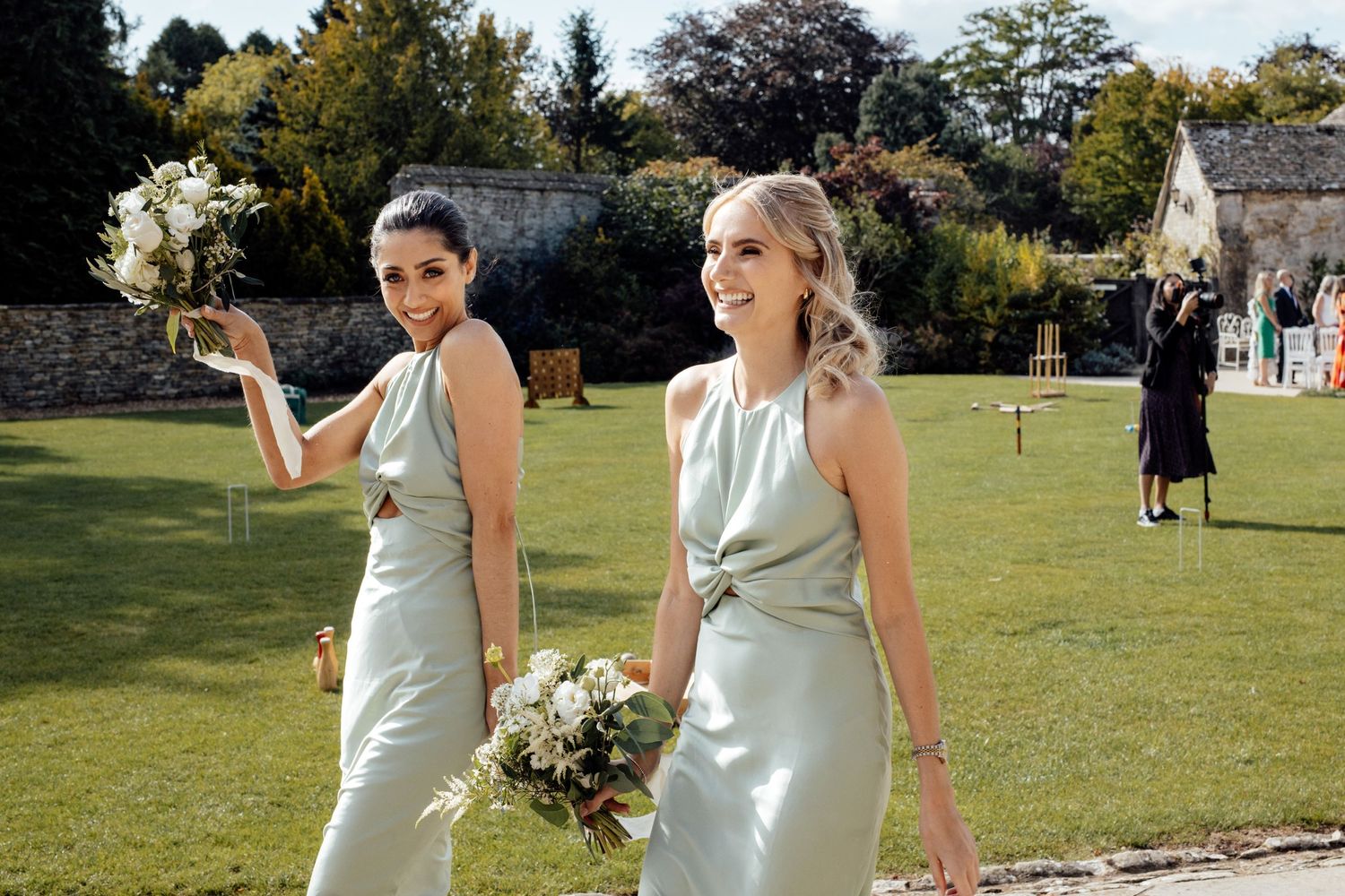 Wedding party members in sage green dresses and grey suits posing outdoors on sunny day.