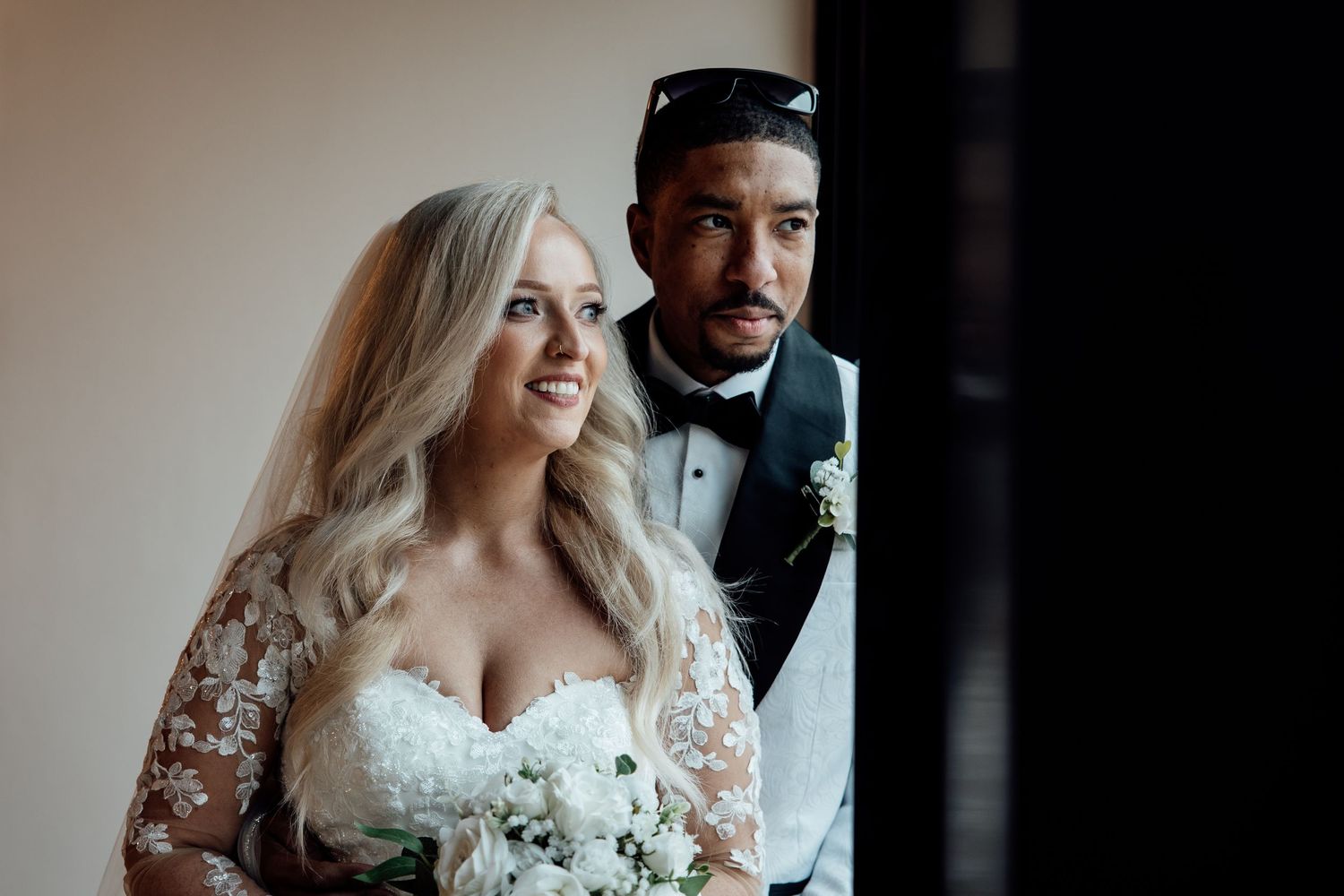 Bride in lace gown holding white bouquet stands with groom in black tuxedo.