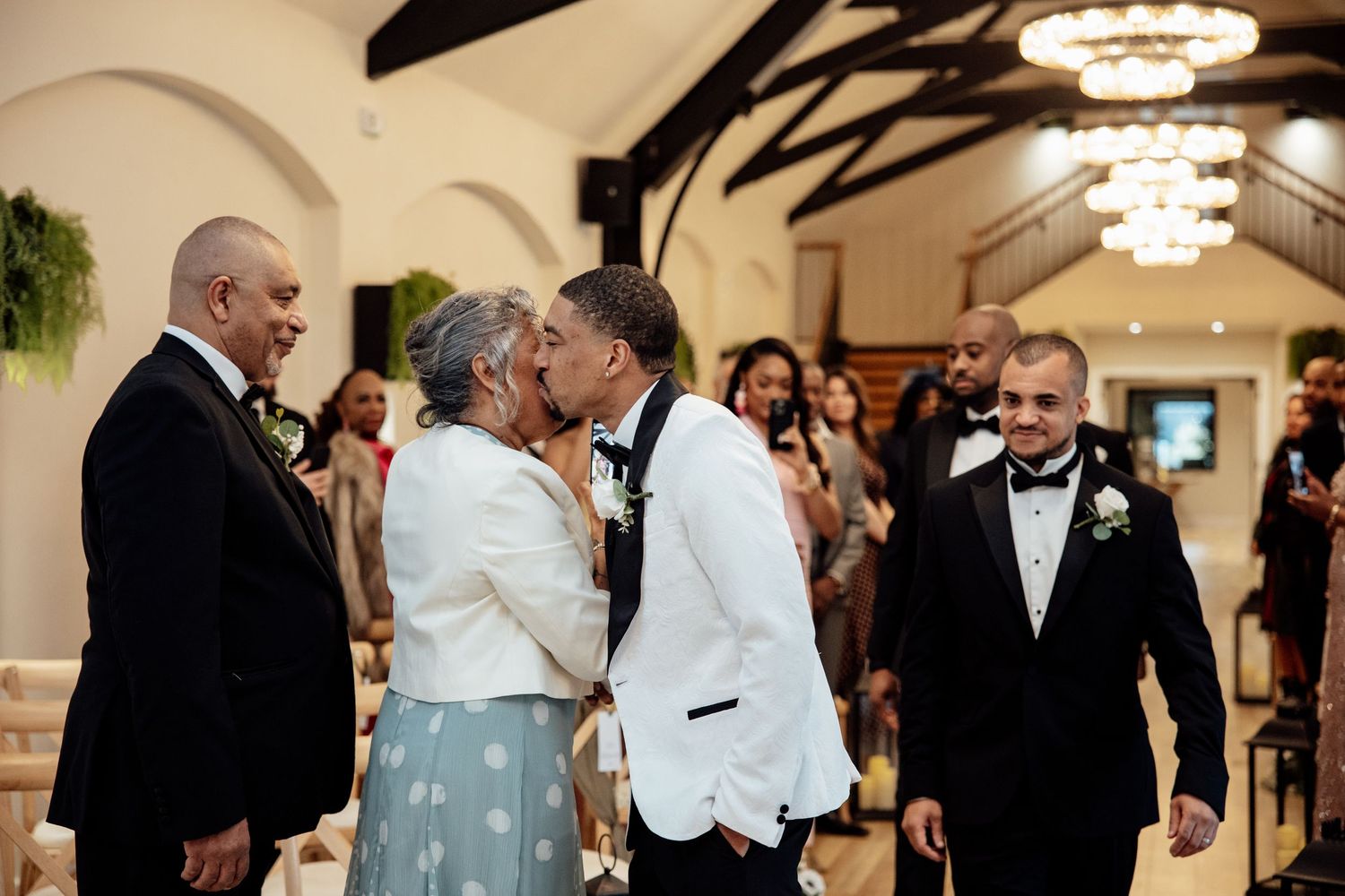 Groom in white tuxedo sharing a kiss with a guest during a joyful indoor wedding ceremony.