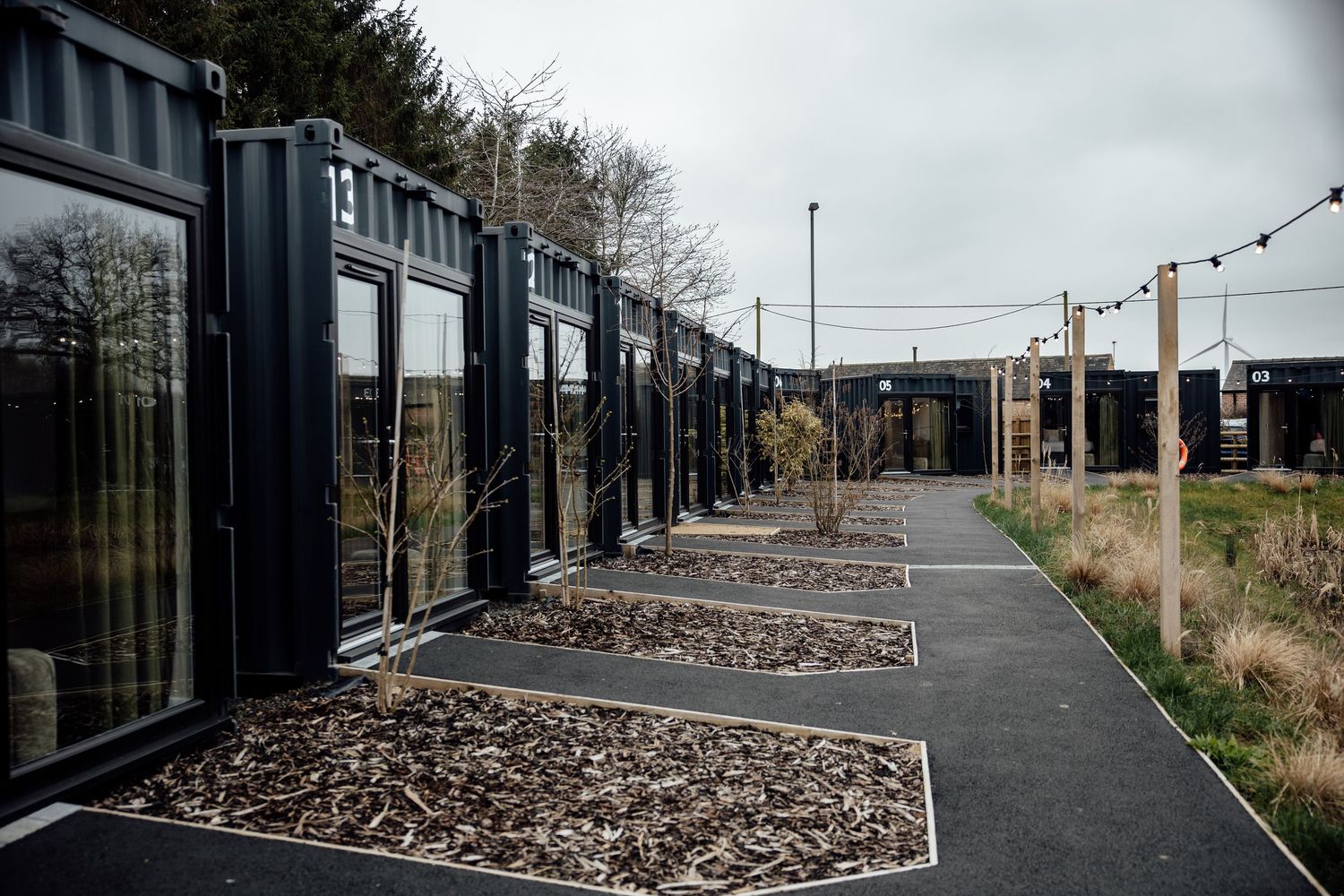 Row of modern black shipping container accommodations with large windows and landscaped mulch beds along a paved pathway.