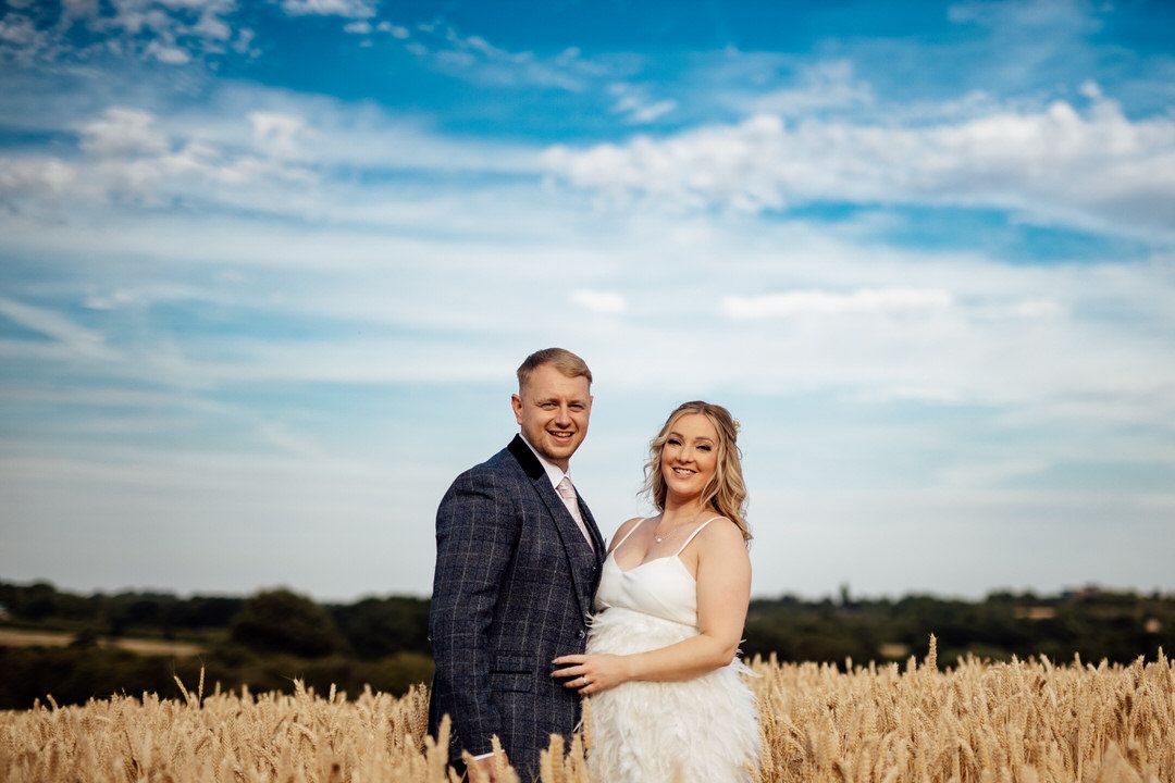 Couple poses in wheat field under bright blue summer sky during wedding photoshoot.