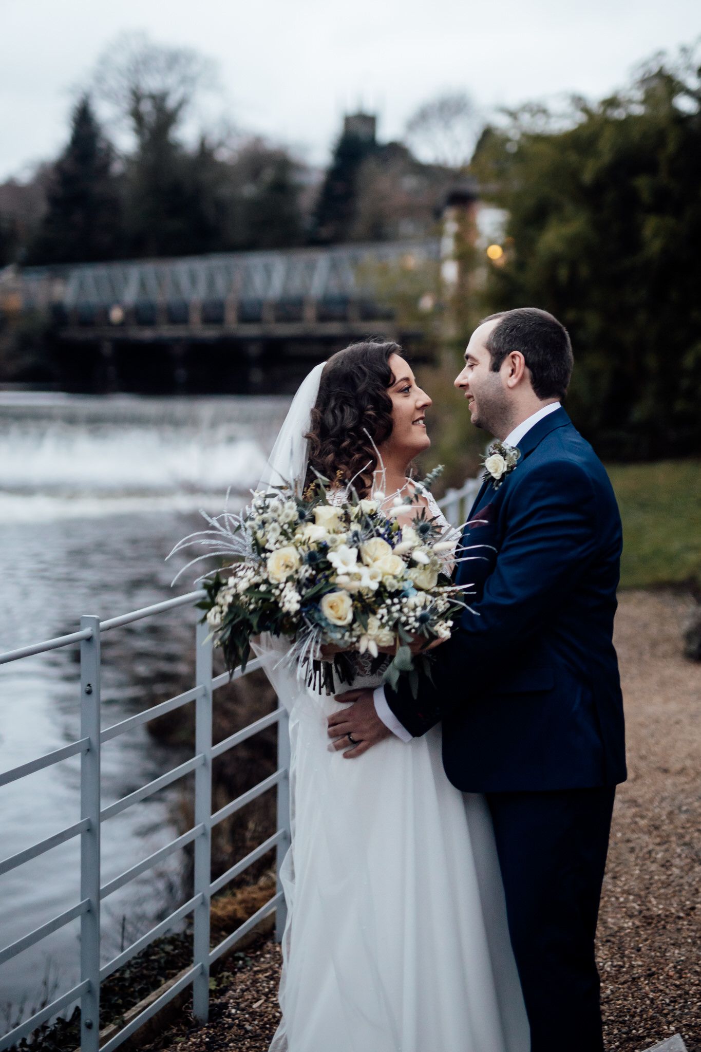 Newlyweds share a romantic moment by the water's edge with winter foliage in the background.